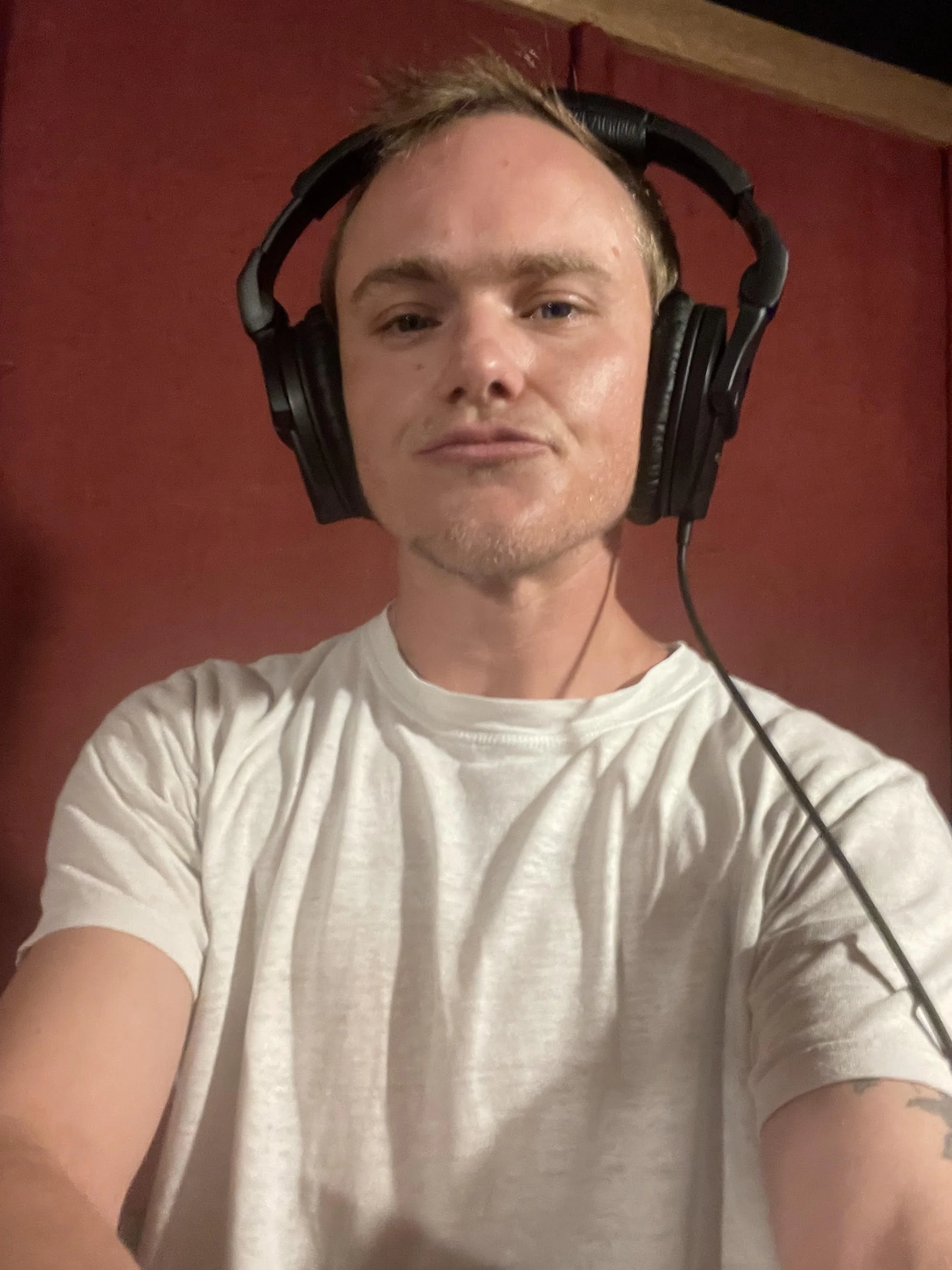 Sandy Ernest Allen, a blonde trans man, in a white shirt, wearing headphones, in a red-painted recording studio