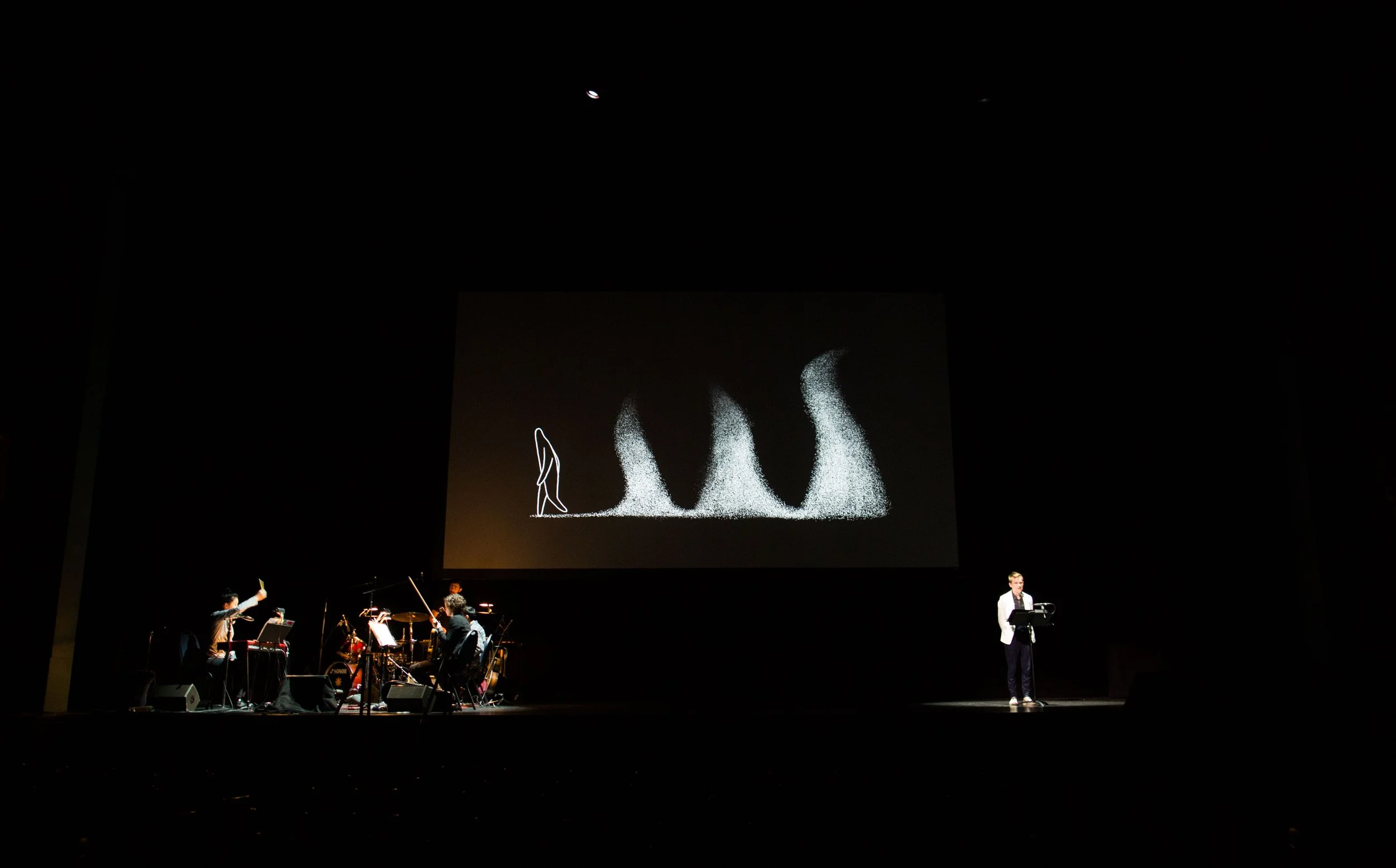 Author Sandy Allen in a white suit jacket on a stage beside a small orchestra with a ghostly illustration projected behind