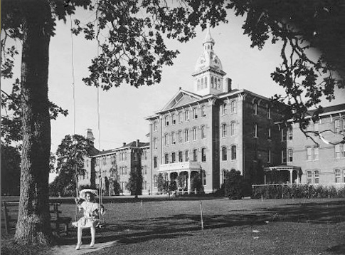 A historic photograph of a Kirkbride asylum with a patient in front on a swing