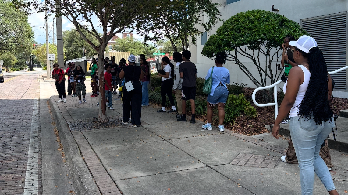 A group of people standing in line outside a building on a sidewalk under trees during the WHAT IT WAS WALKING TOUR in Tampa.