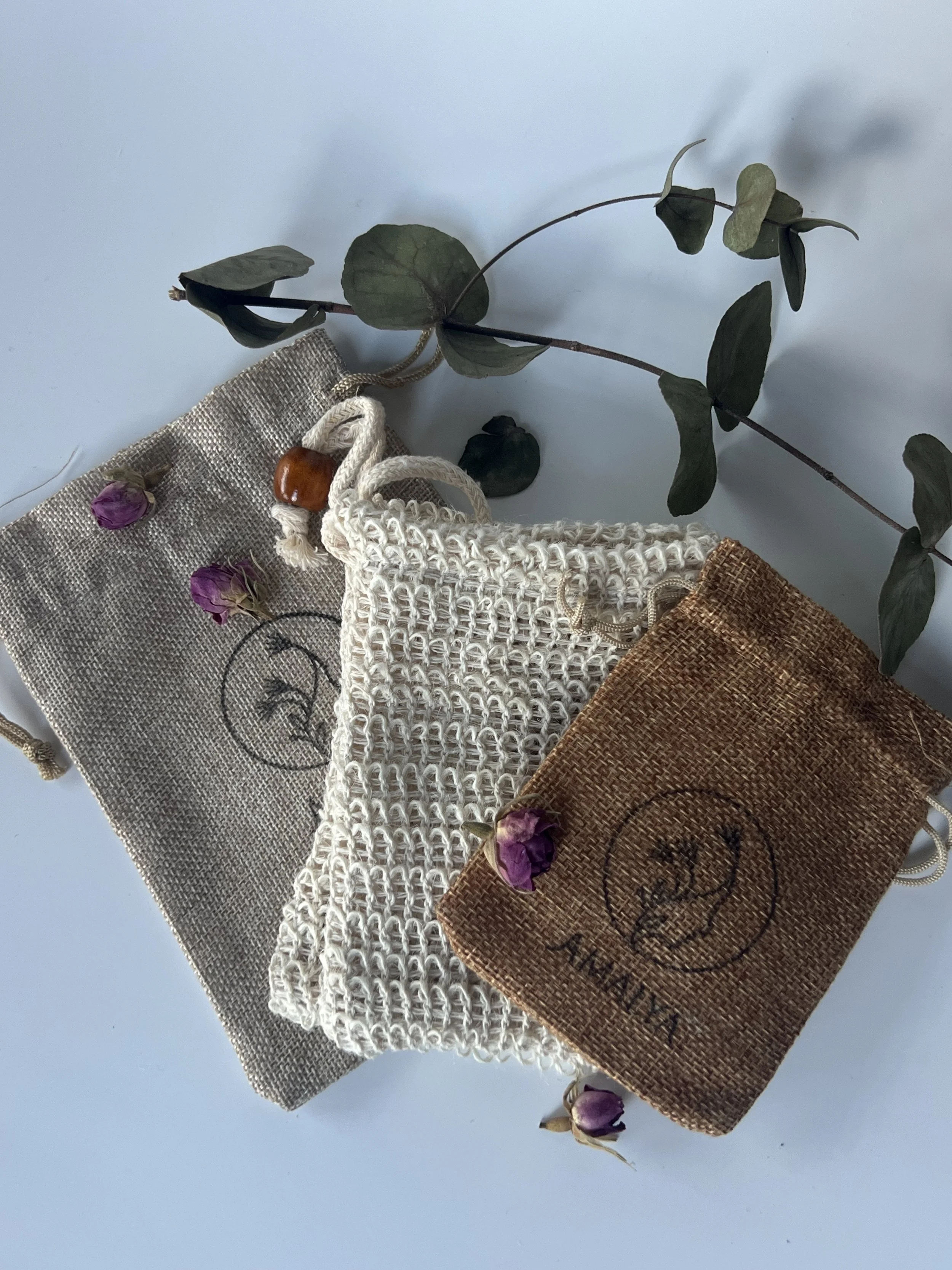 Three fabric pouches, one grey, one white crocheted, and one brown, with a branch of green leaves and small purple flowers on a light background.