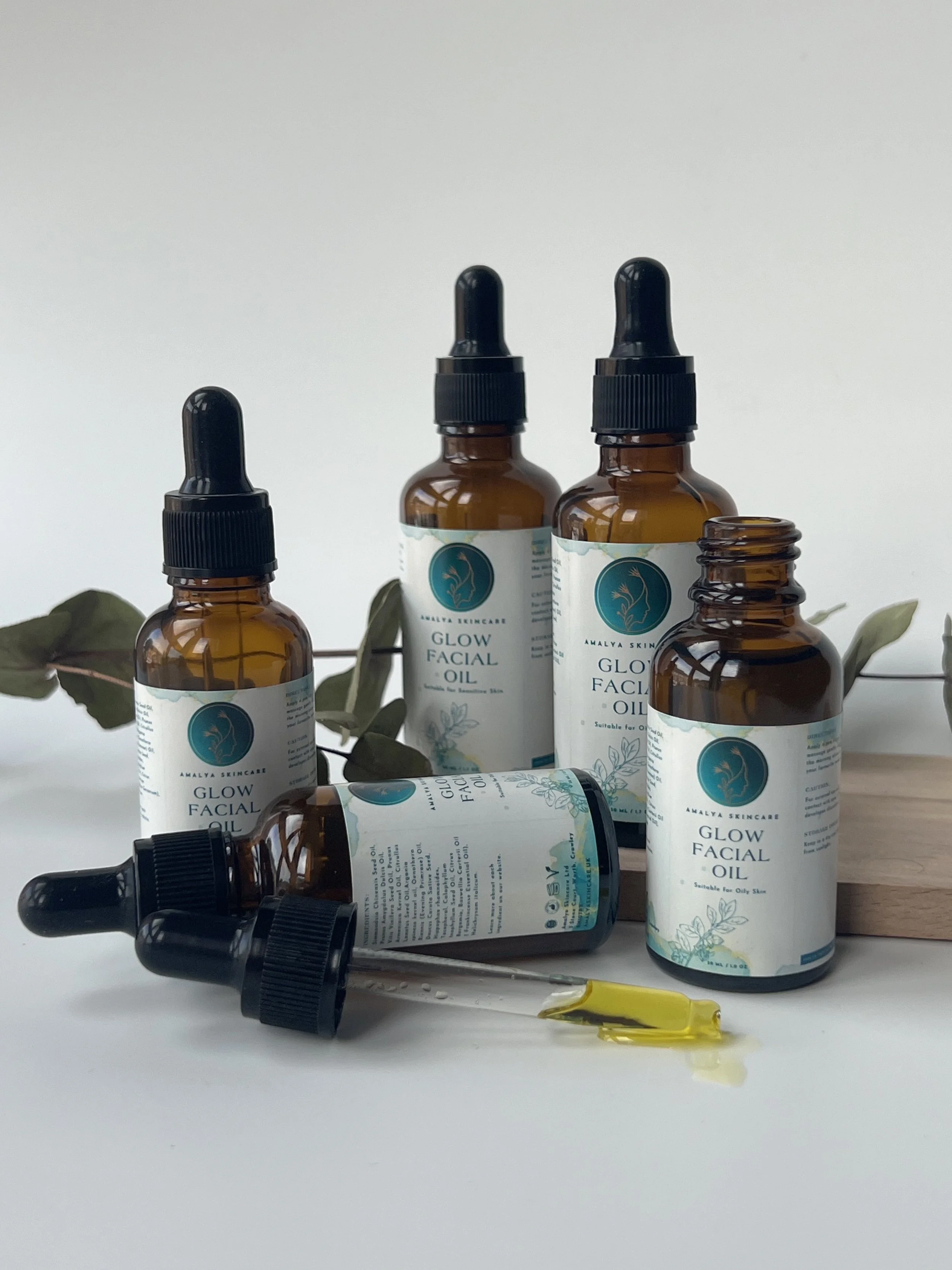 Bottles of skincare facial oil with droppers, labeled "Glow Facial Oil," on a white surface with leaves in the background.