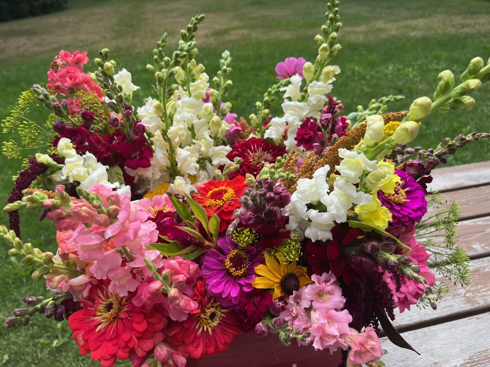 Colorful bouquet of various flowers including pink, red, white, yellow, and purple blooms, arranged on a wooden bench outdoors.