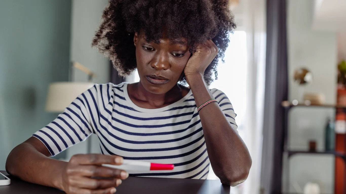 A woman with curly hair looking sad or worried while holding a thermometer. She is sitting at a table in a room with a lamp and shelves in the background.