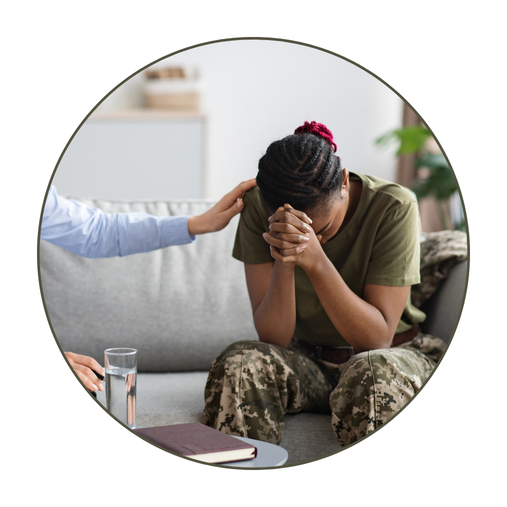 A woman in military camouflage clothing sitting on a sofa with her head bowed and hands clasped in front of her face, while a person beside her offers comfort by touching her shoulder, in a living room setting.