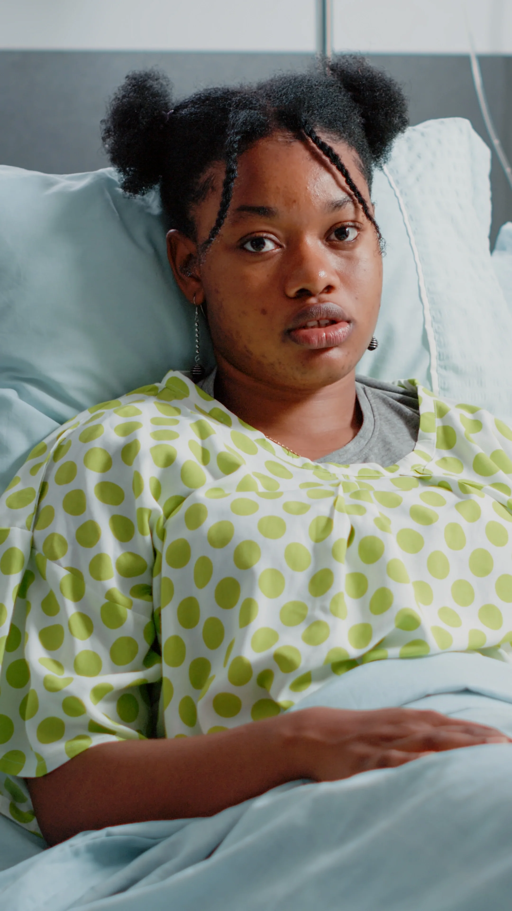 Young woman lying in hospital bed, wearing a green polka-dot hospital gown, looking at the camera, with a neutral expression.