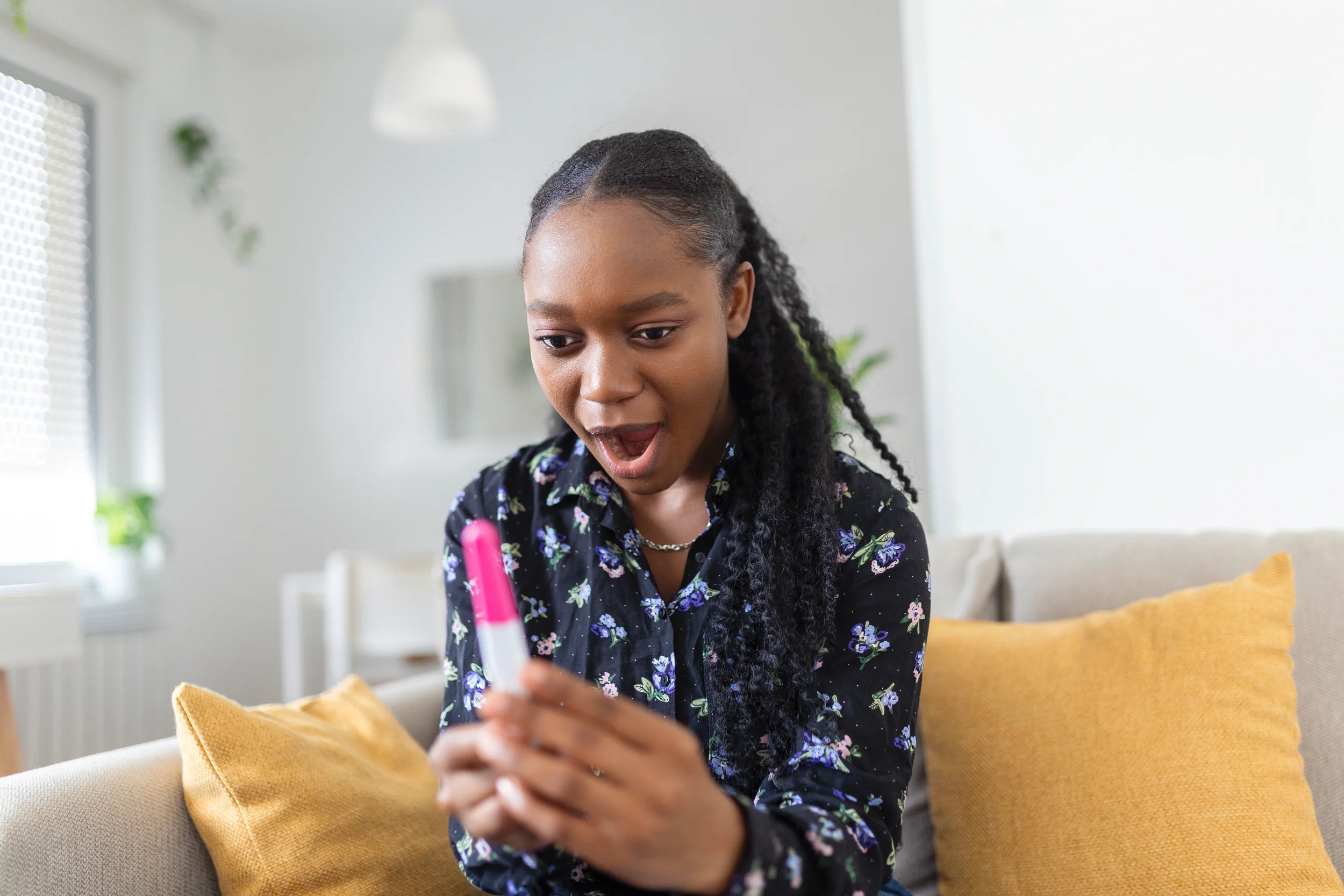 A young woman with braided hair wearing a dark floral shirt, sitting on a beige sofa with a yellow pillow, looking at and reacting to her phone in a bright, modern living room.