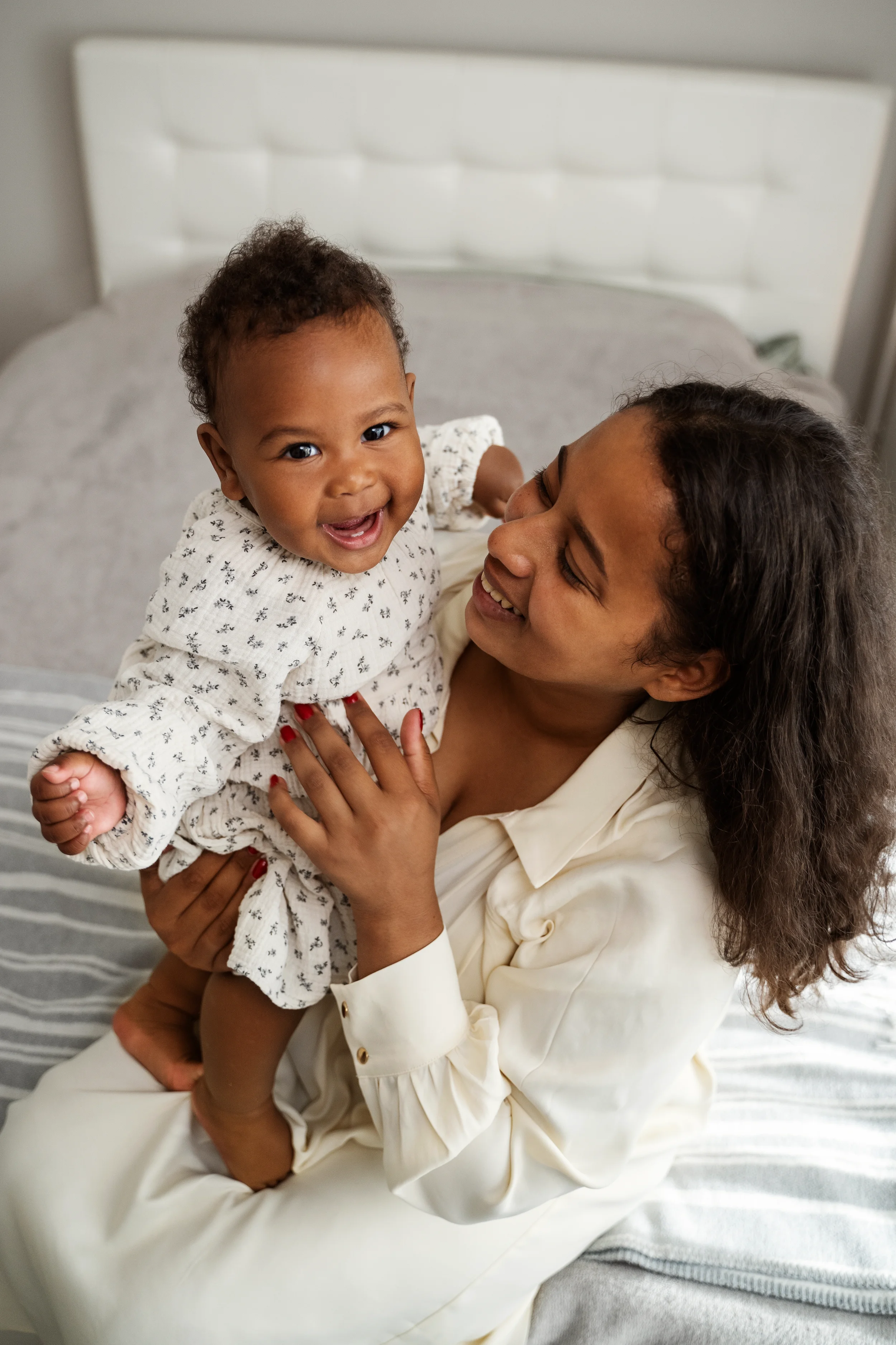 A woman with long brown hair holding a smiling toddler in her arms on a bed with gray and white striped sheets.