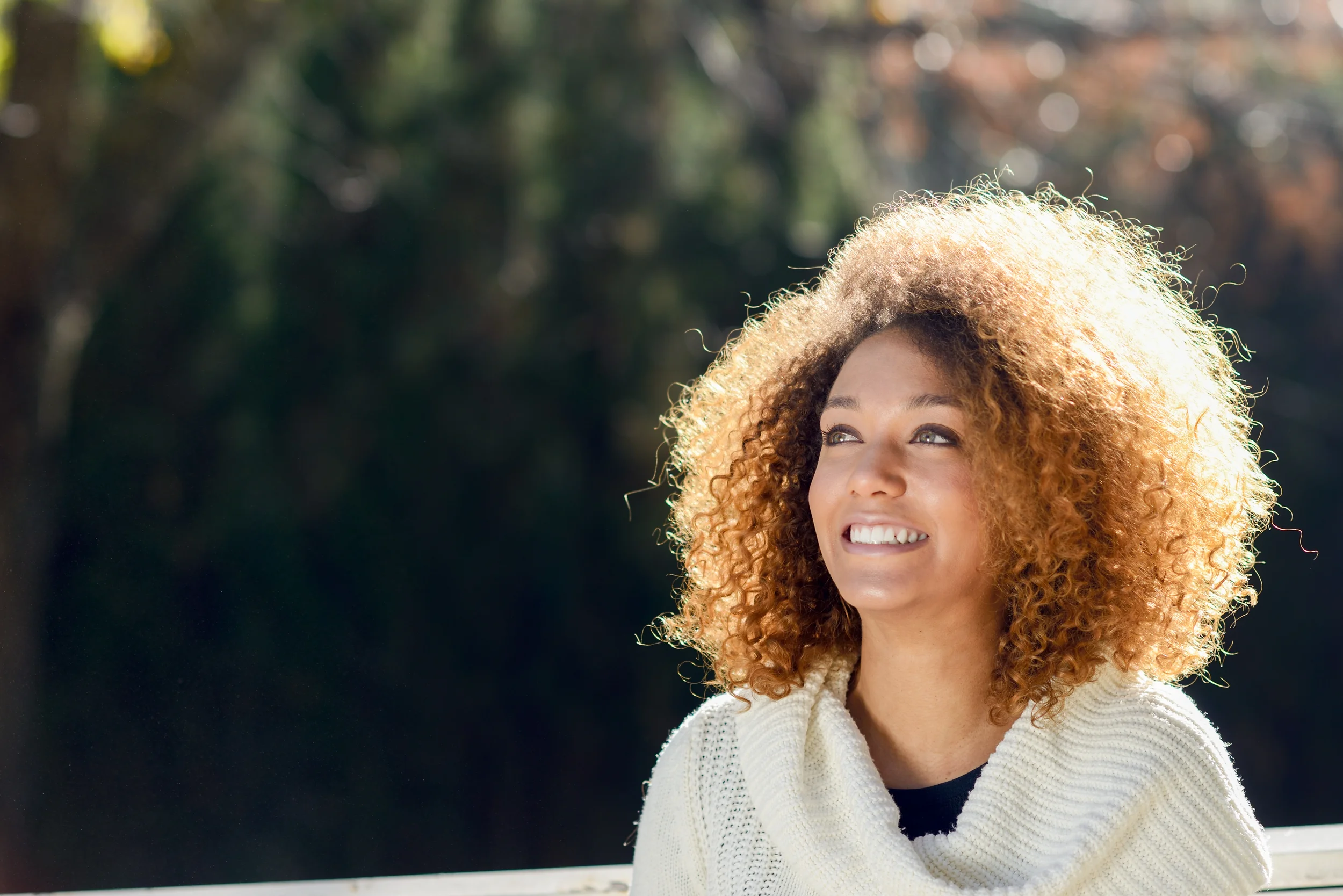 A woman with curly blonde hair smiling outdoors on a sunny day, wearing a white knitted sweater.