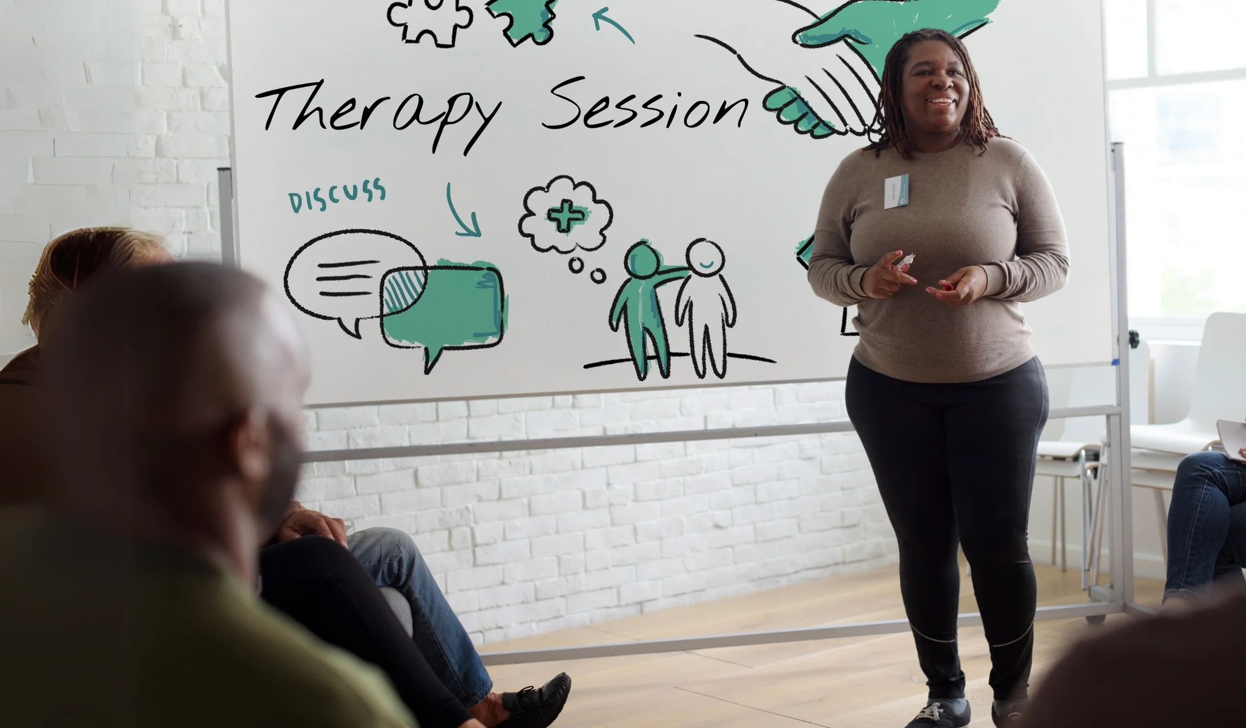 A woman giving a presentation about therapy session in a room with a whiteboard, and a few attendees seated listening.