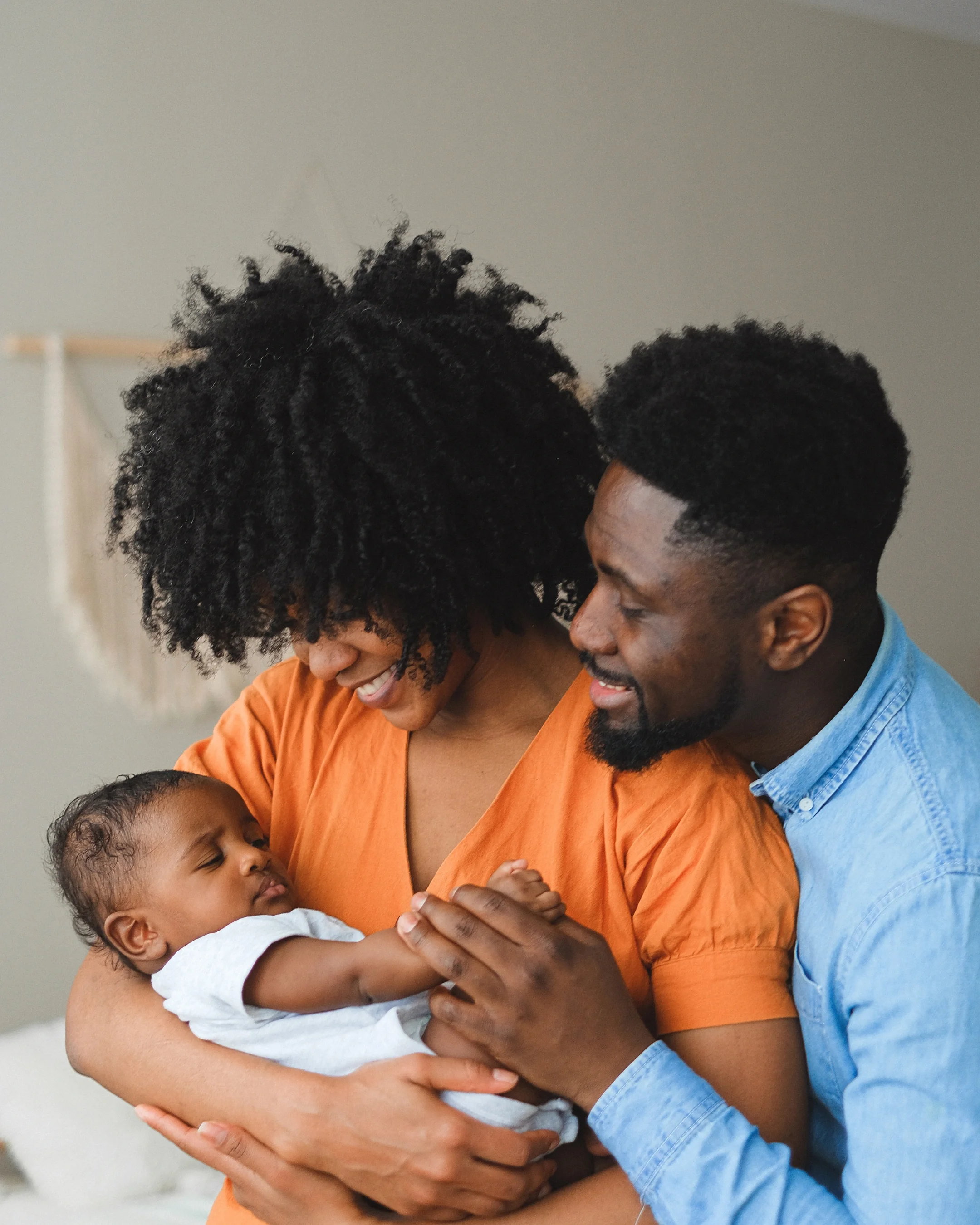 A happy family of three, with a mother holding an infant and a father affectionately touching the baby, all smiling in a cozy indoor setting.