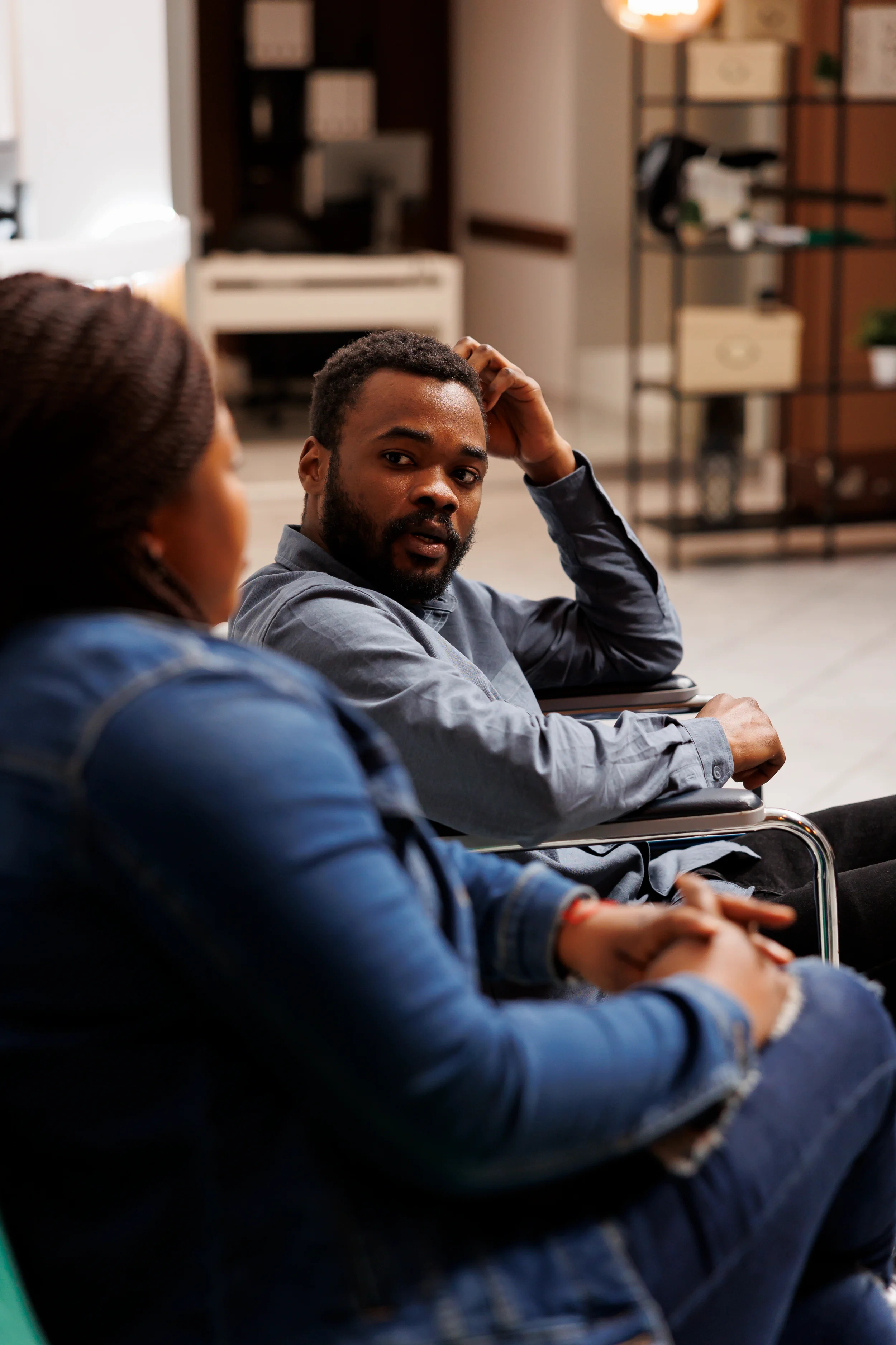 A man and woman sitting on chairs in a room, having a conversation. The man looks curious or concerned, while the woman listens attentively.