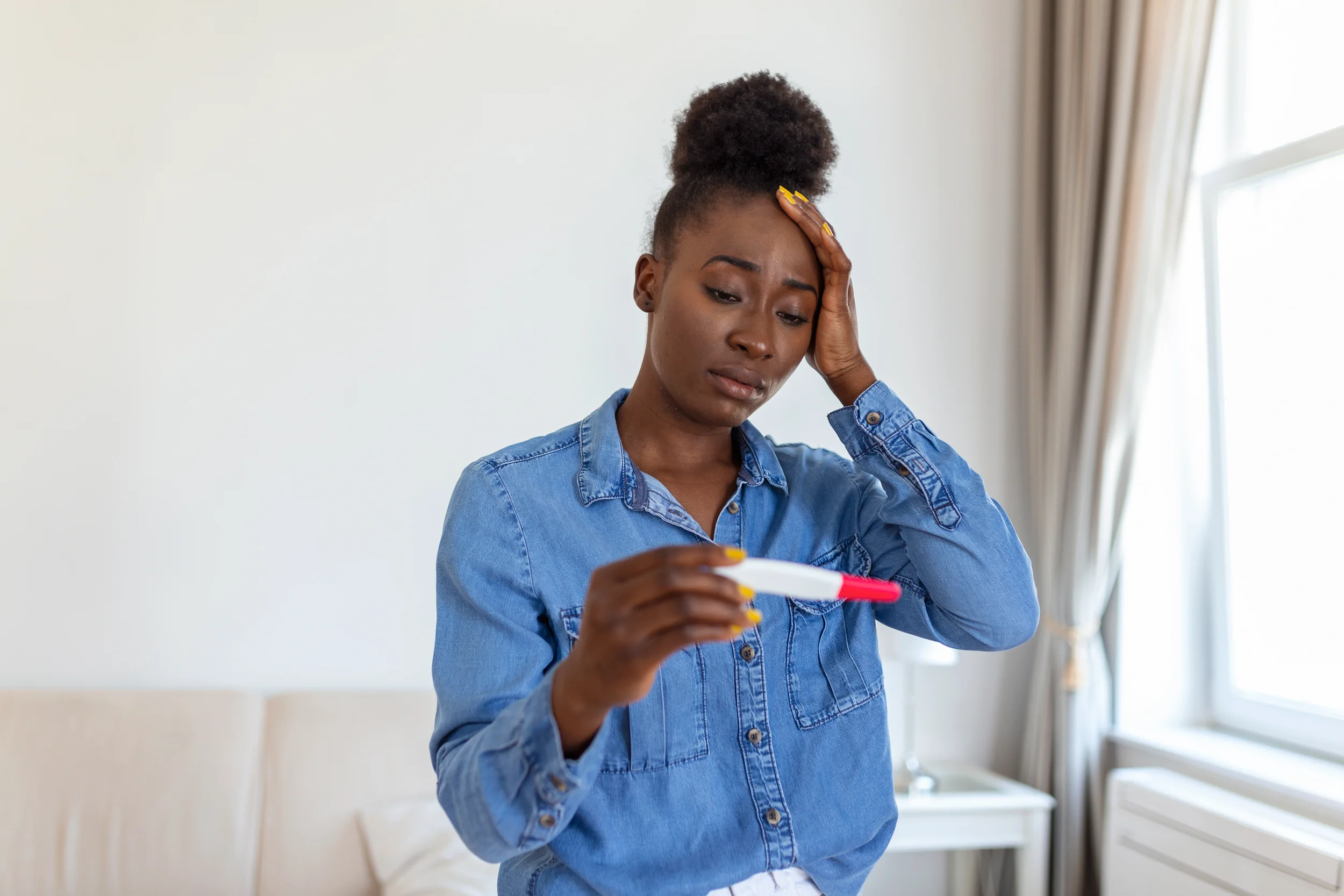 Young woman with curly hair in a bun, wearing a blue denim shirt, looks worried while holding a pregnancy test in her hand.