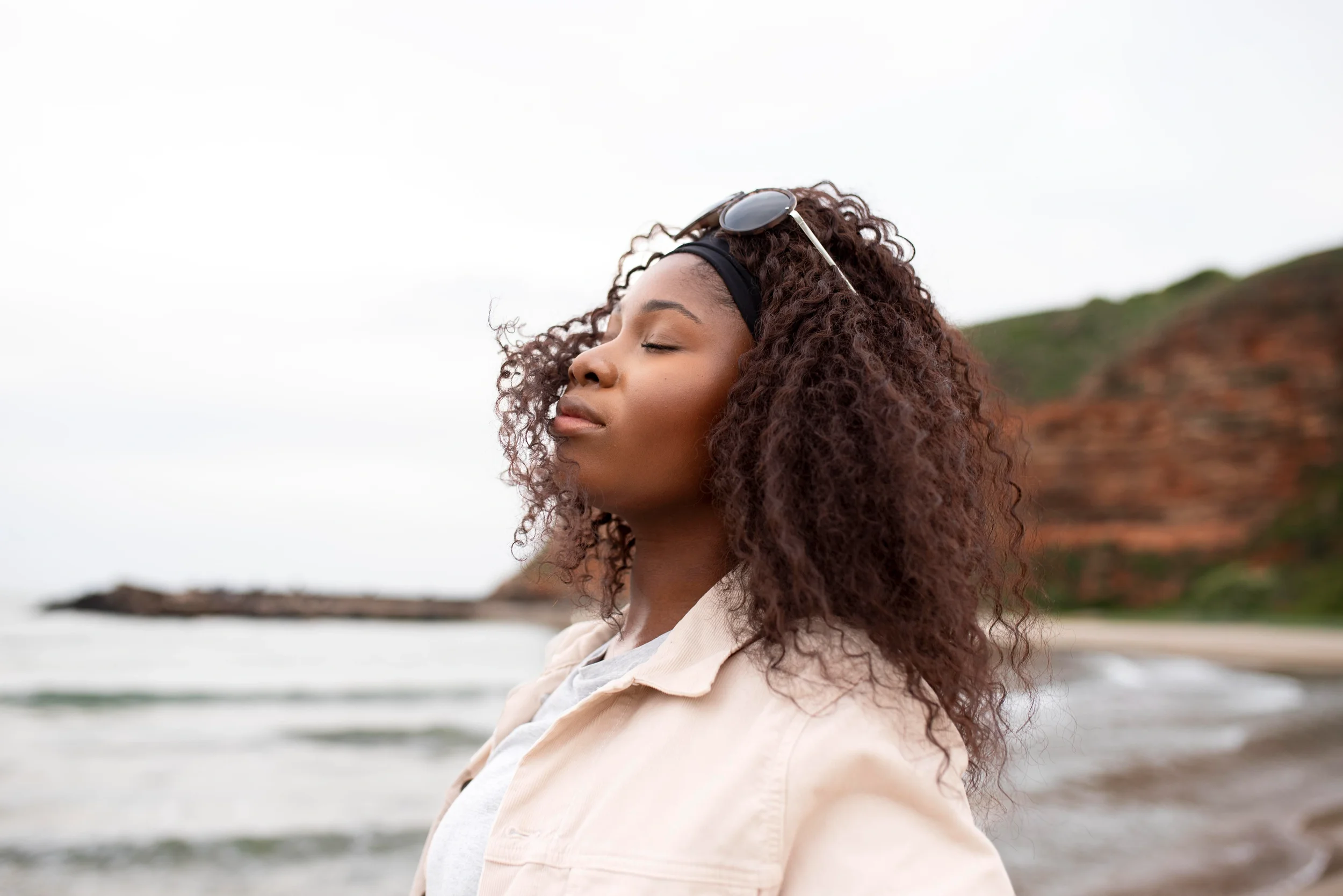 Young woman with curly hair and sunglasses resting on her head, closing her eyes at the beach with cliffs in the background.