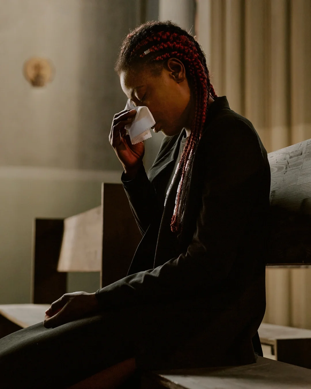 A woman with red braids sitting on a bench in a church, holding a tissue to her face, appearing emotional or crying.