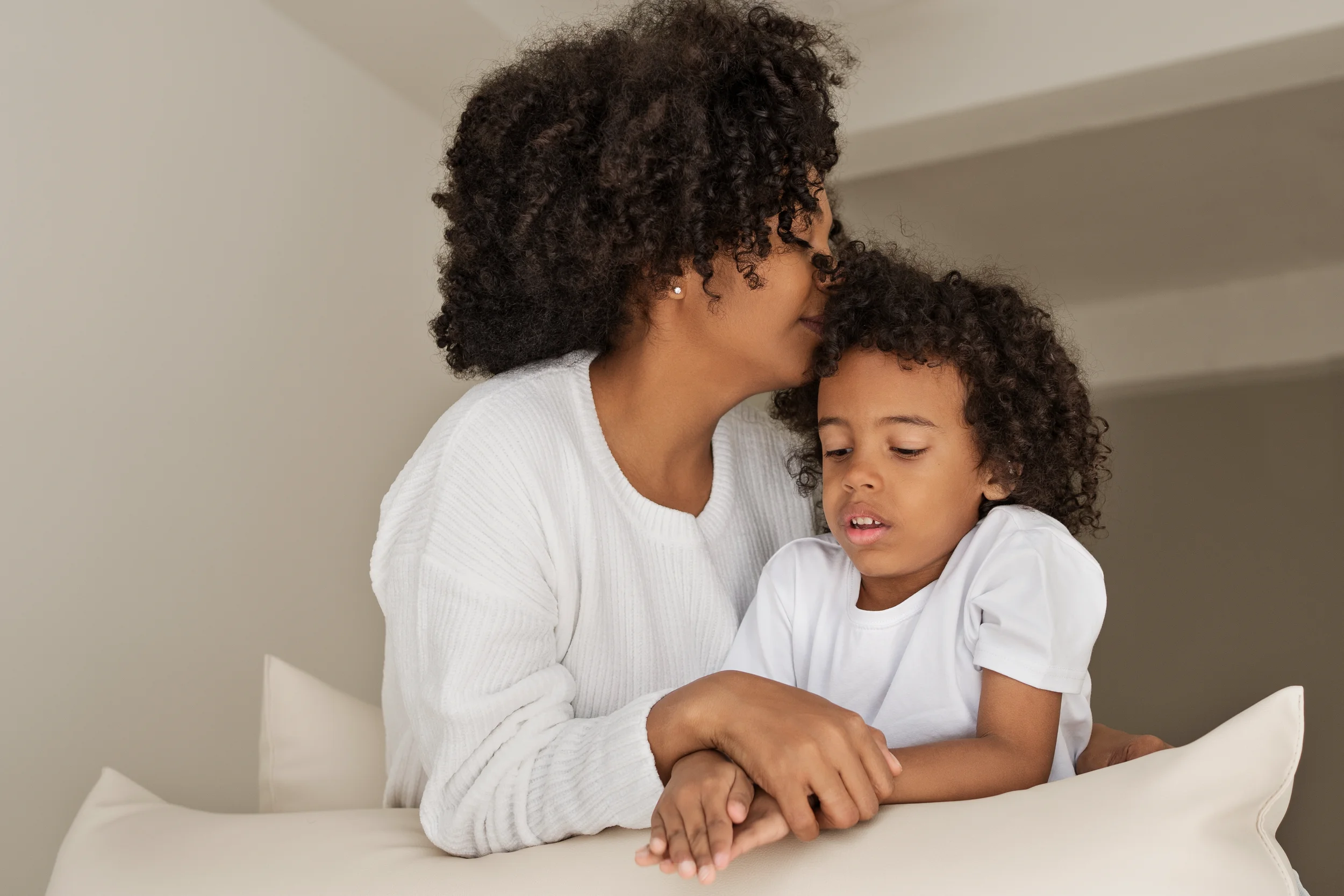 A woman with curly hair and earrings gently kisses a young girl's forehead while holding her hands, both sitting at a table in a neutral-colored room.