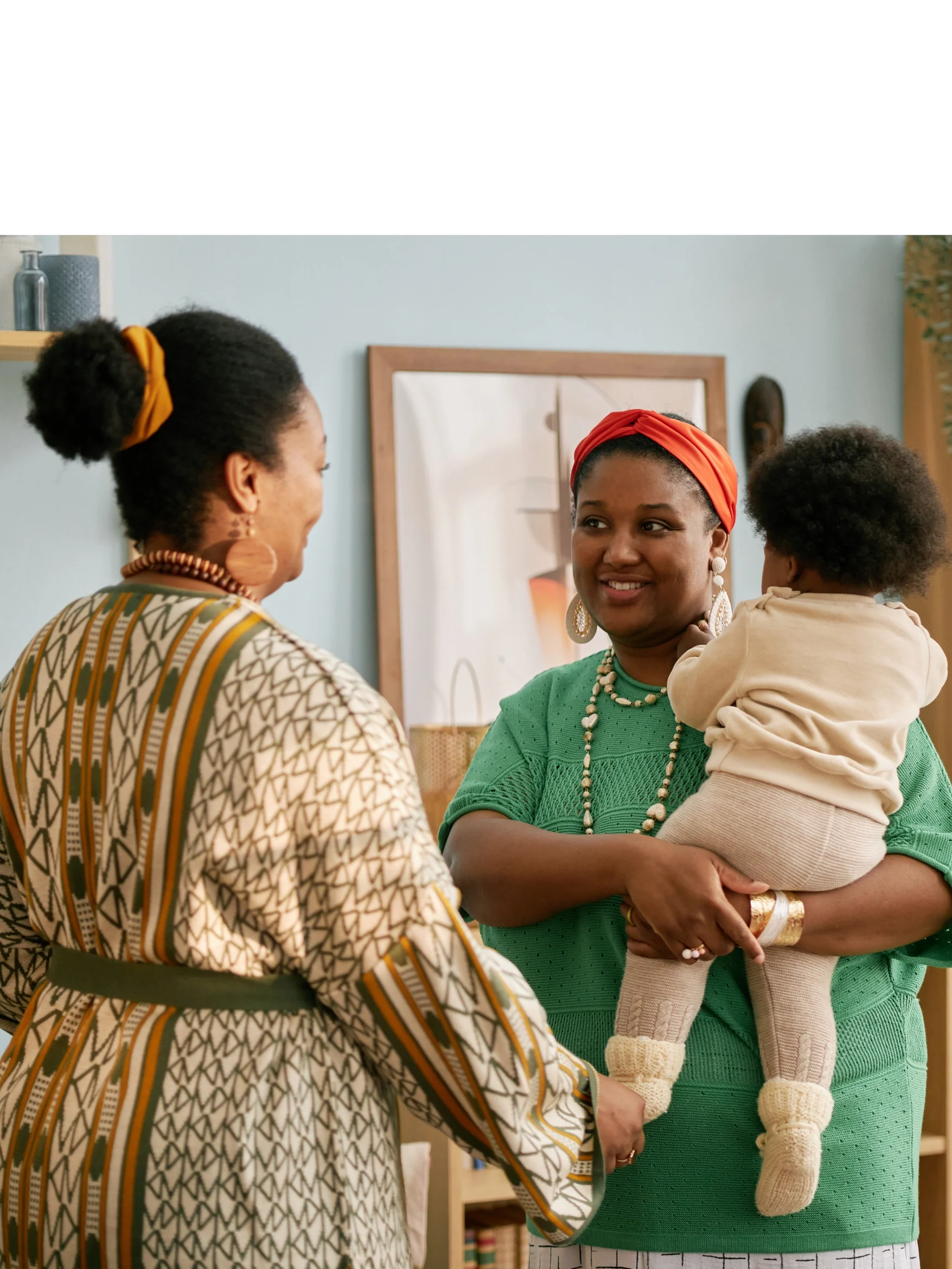 Two women talking indoors, one holding a young child, with a framed picture on the wall behind them.