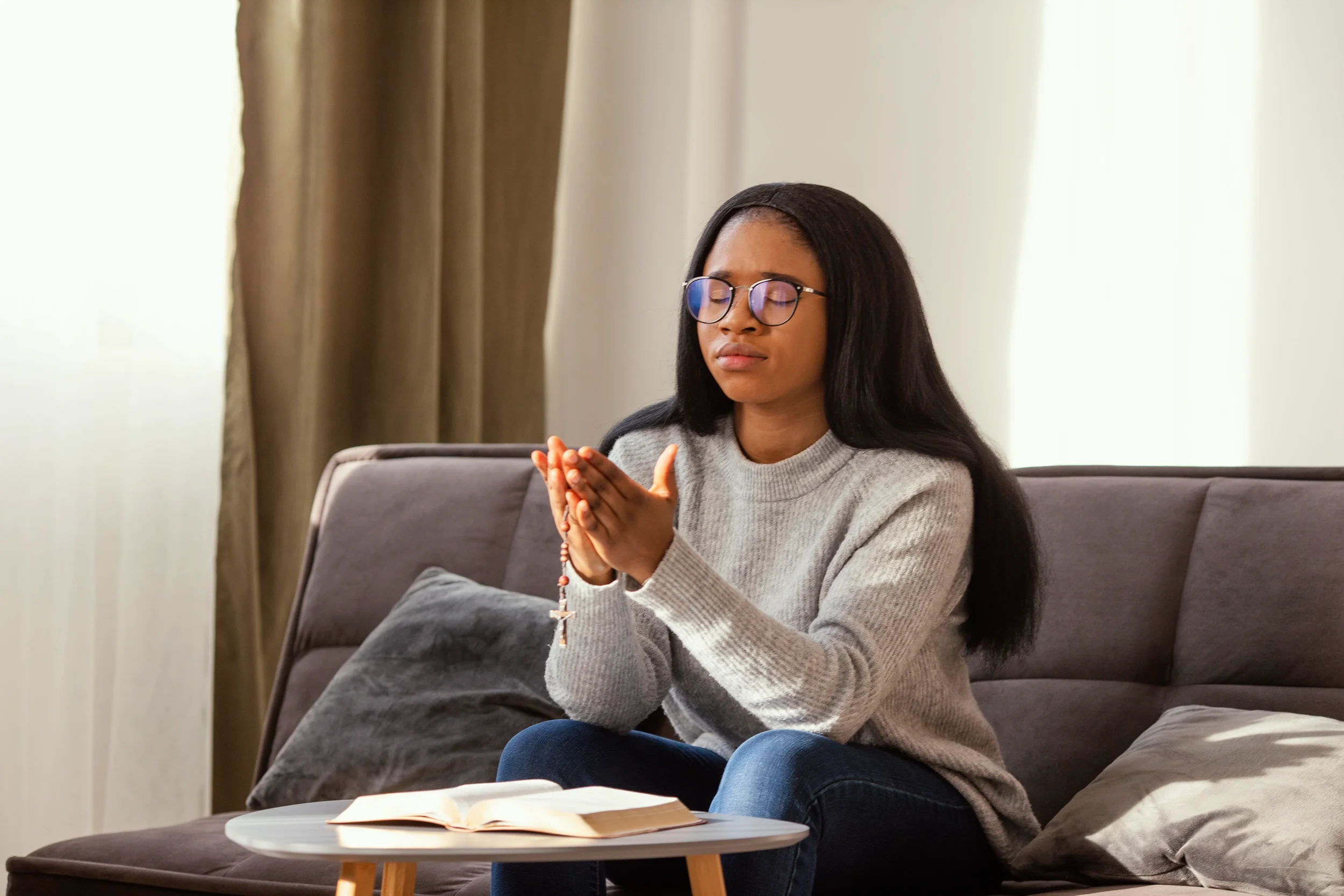 A young woman with glasses praying with a rosary, sitting on a sofa with an open book and a table in front of her.