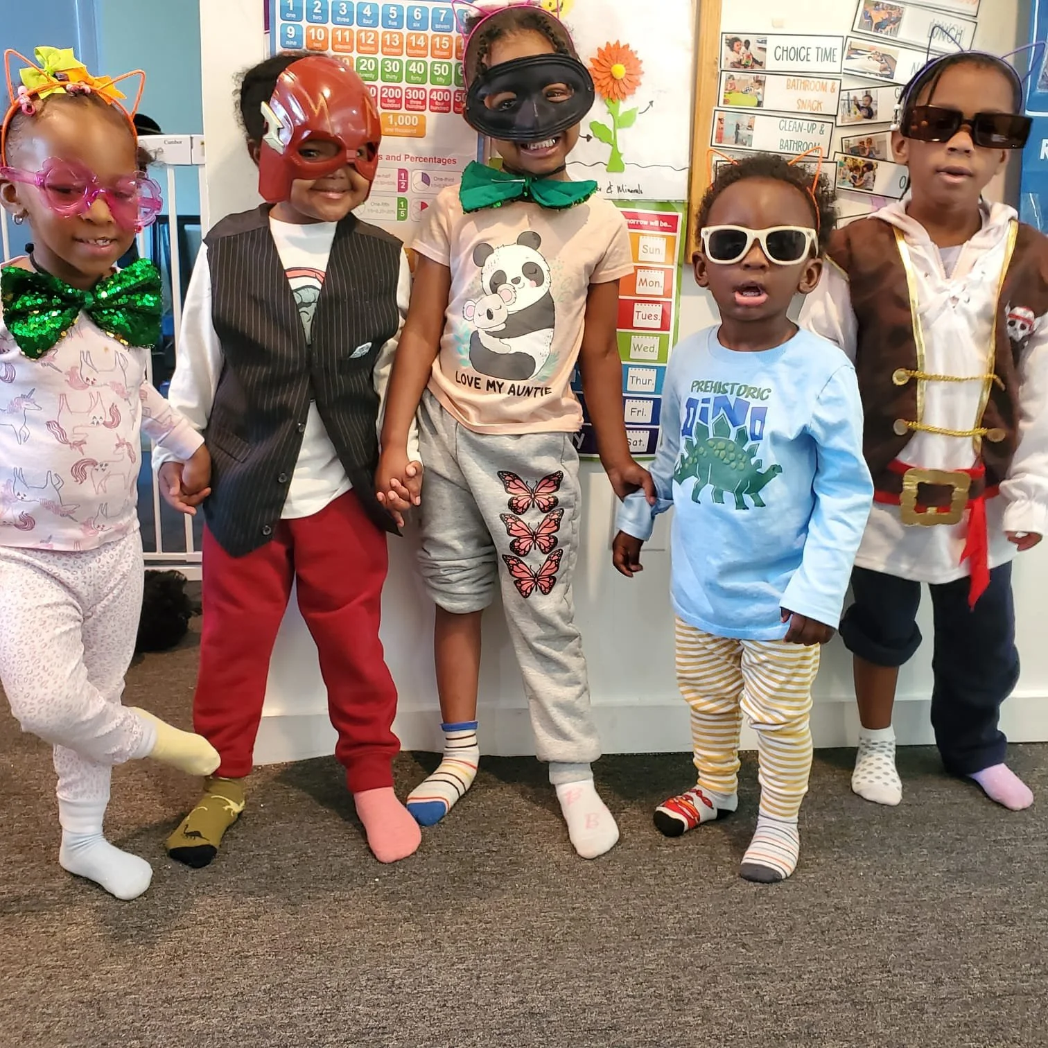 Children dressed in colorful costumes with hats, glasses, and socks standing in a row in a classroom with educational charts and a schedule in the background.