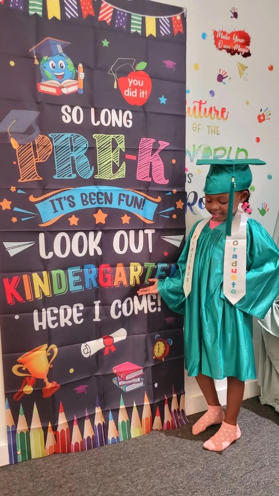 A young girl in a teal graduation gown and cap standing in front of a colorful kindergarten graduation sign. The sign features various bright colors, congratulatory messages, and school-themed illustrations, celebrating her transition to elementary s