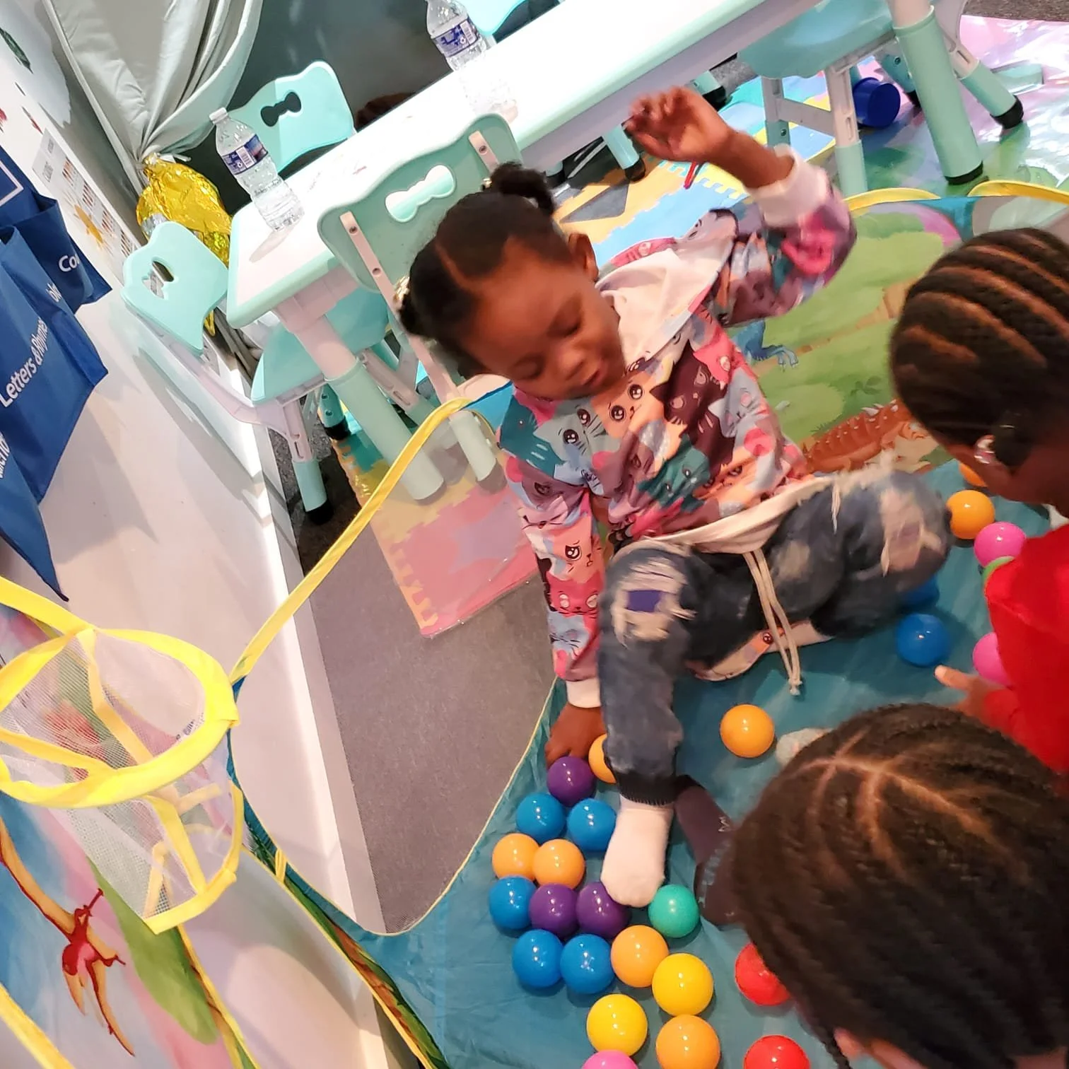 Children playing a ball toss game at an indoor party or event, with colorful plastic balls and a mesh basket nearby.