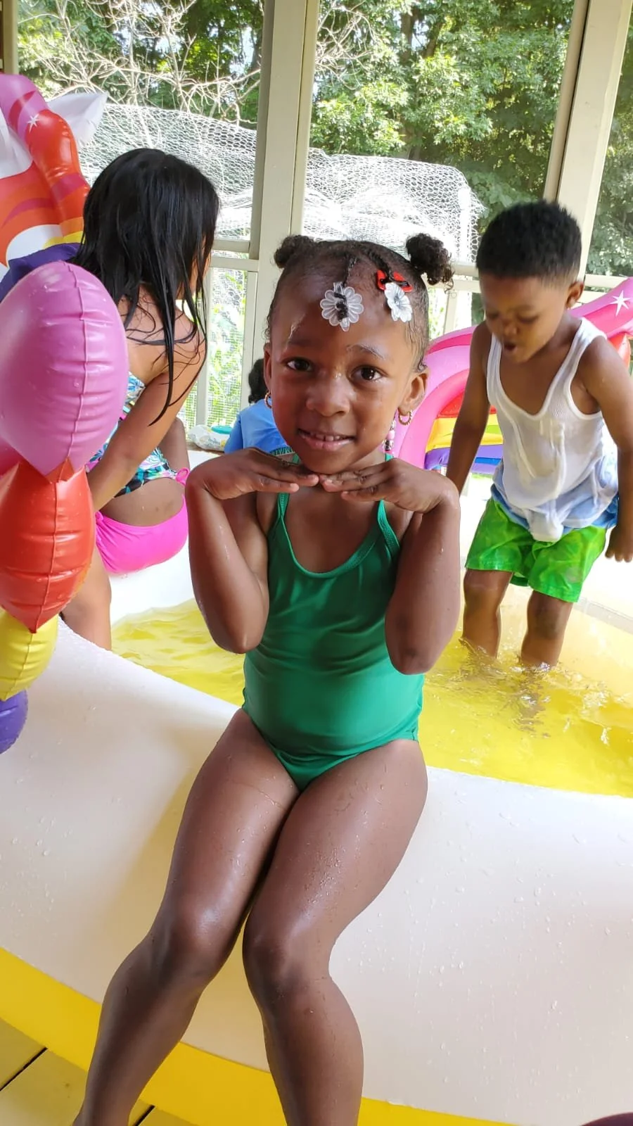 Young girl in a green swimsuit sitting on the edge of a small pool, smiling with hands under her chin, surrounded by other children playing in the pool with inflatable toys and water, indoors with a screened window and trees outside.