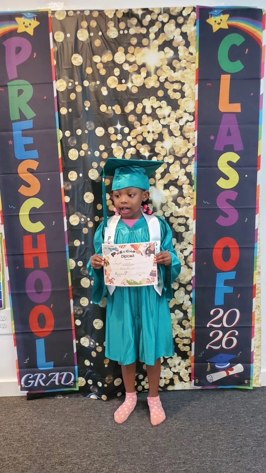 Young girl in teal graduation gown and cap holding diploma standing in front of a celebratory backdrop with the words 'PreSchool' and 'Class of 2026', decorated with colorful banners, stars, and a rainbow.