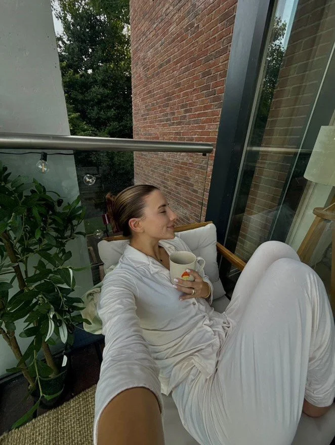 Woman relaxing on a balcony, smiling, holding a mug, wearing cream pajamas, with a large plant and brick wall in background.