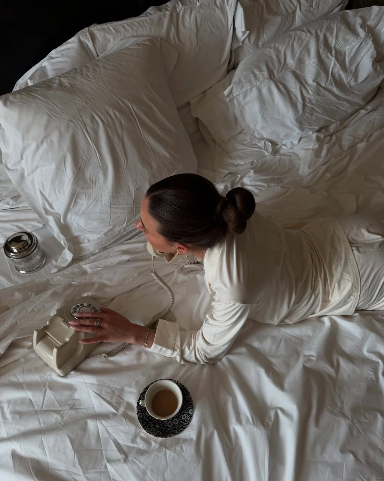 A woman with dark hair tied in a bun, dressed in buttery soft luxurious pajamas, lying on a bed with white rumpled sheets, talking on a vintage beige rotary phone, with a cup of coffee on a black and white saucer nearby.