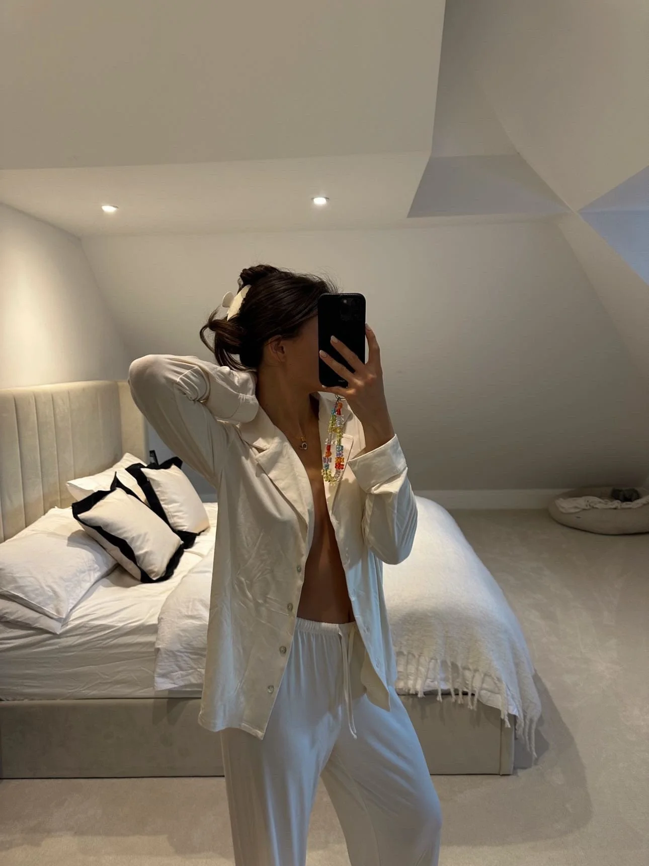 A woman taking a mirror selfie in an elegant bedroom, dressed in a cream luxurious chic pajama set, with a hair clip and necklace, standing in front of a bed with white sheets and black-and-white pillows.