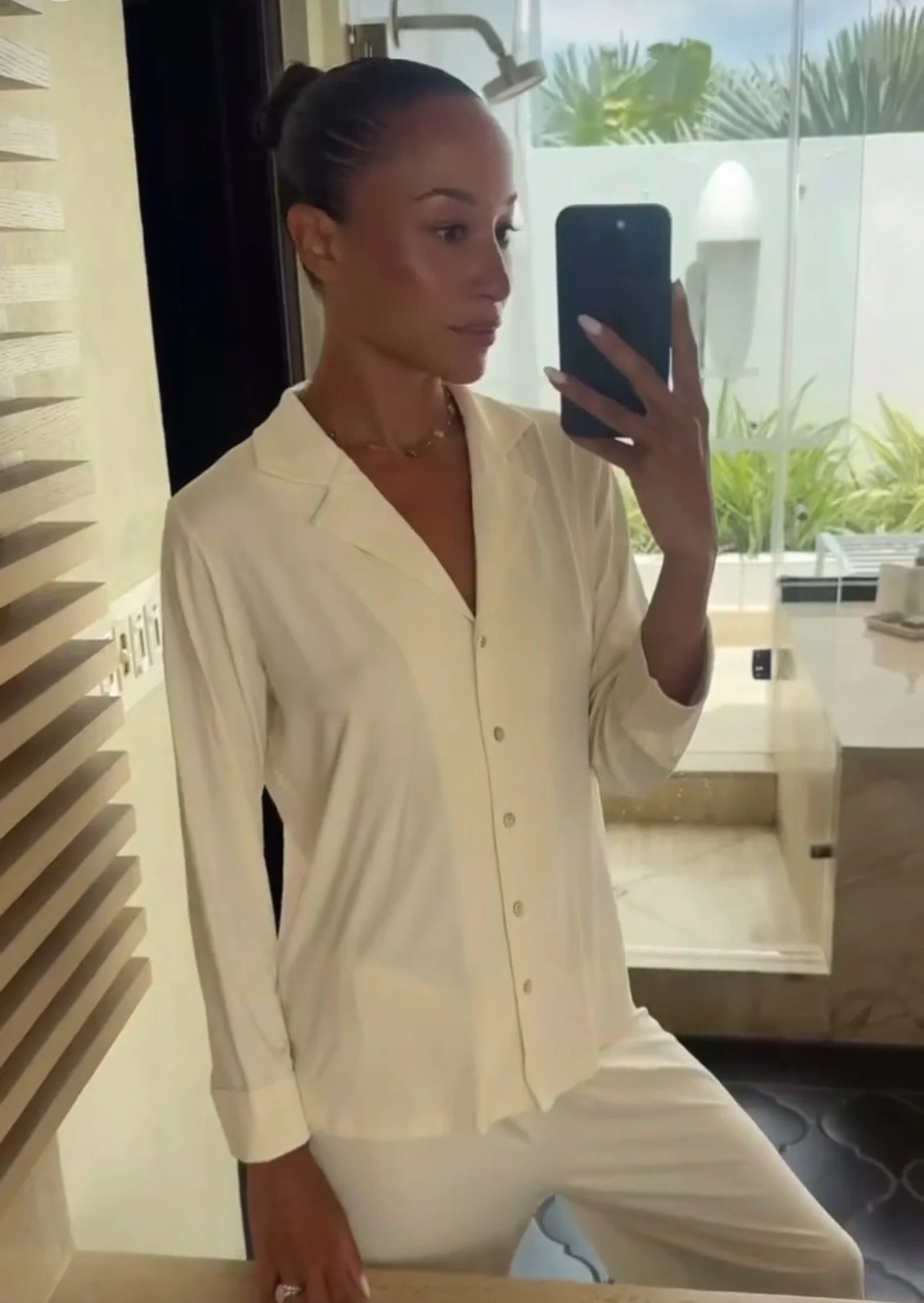 Woman taking a mirror selfie in a bright room with greenery outside, wearing a cream-colored button-down shirt and matching pants.