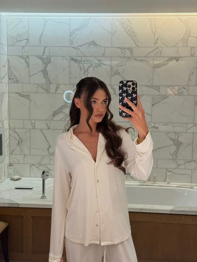 A woman taking a mirror selfie in a bathroom. She has long dark hair in loose waves, wearing a cream-colored luxurious pajama top and matching pants set. The bathroom features marble walls and a bathtub in the background.