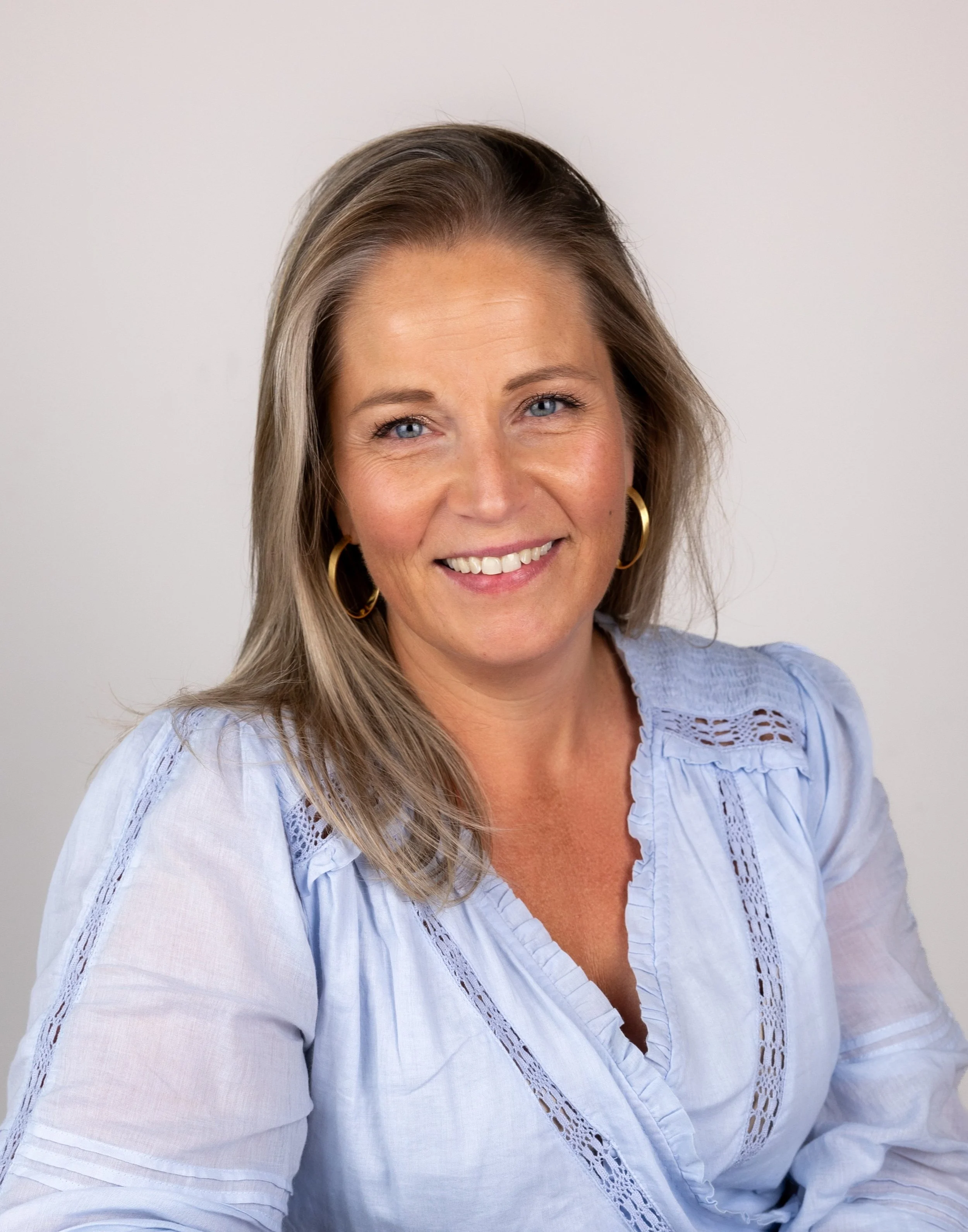 Portret van een lachende vrouw met blond haar, blauwe ogen, grote goudkleurige oorbellen en een lichte blauwe blouse voor een witte achtergrond.