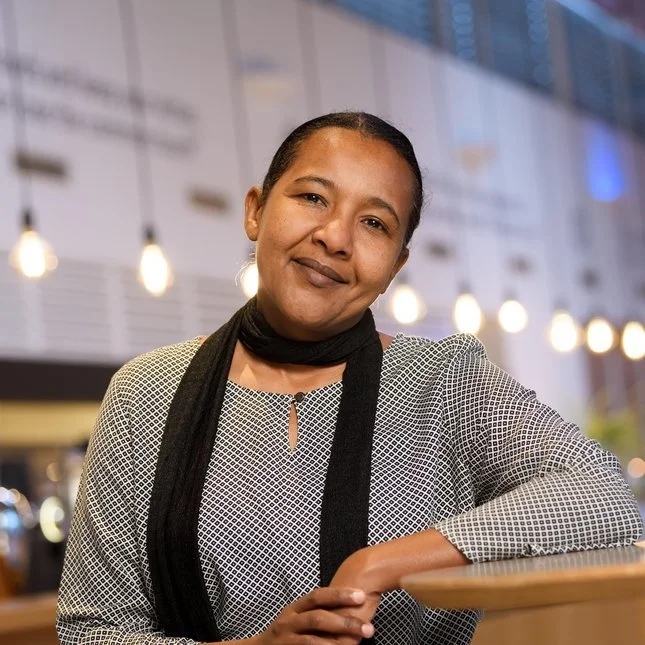 Vrouw met een zwarte sjaal en witte blouse in een modern interieur met hangende lampen.