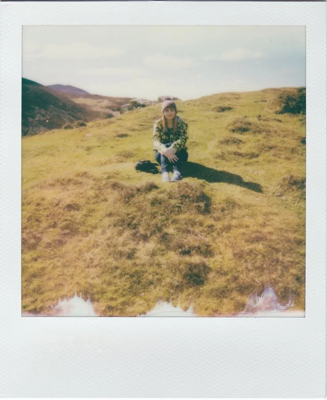 Polaroid of a woman sitting on a grassy hillside in soft sunlight, shared anonymously as part of an ED recovery quote series.