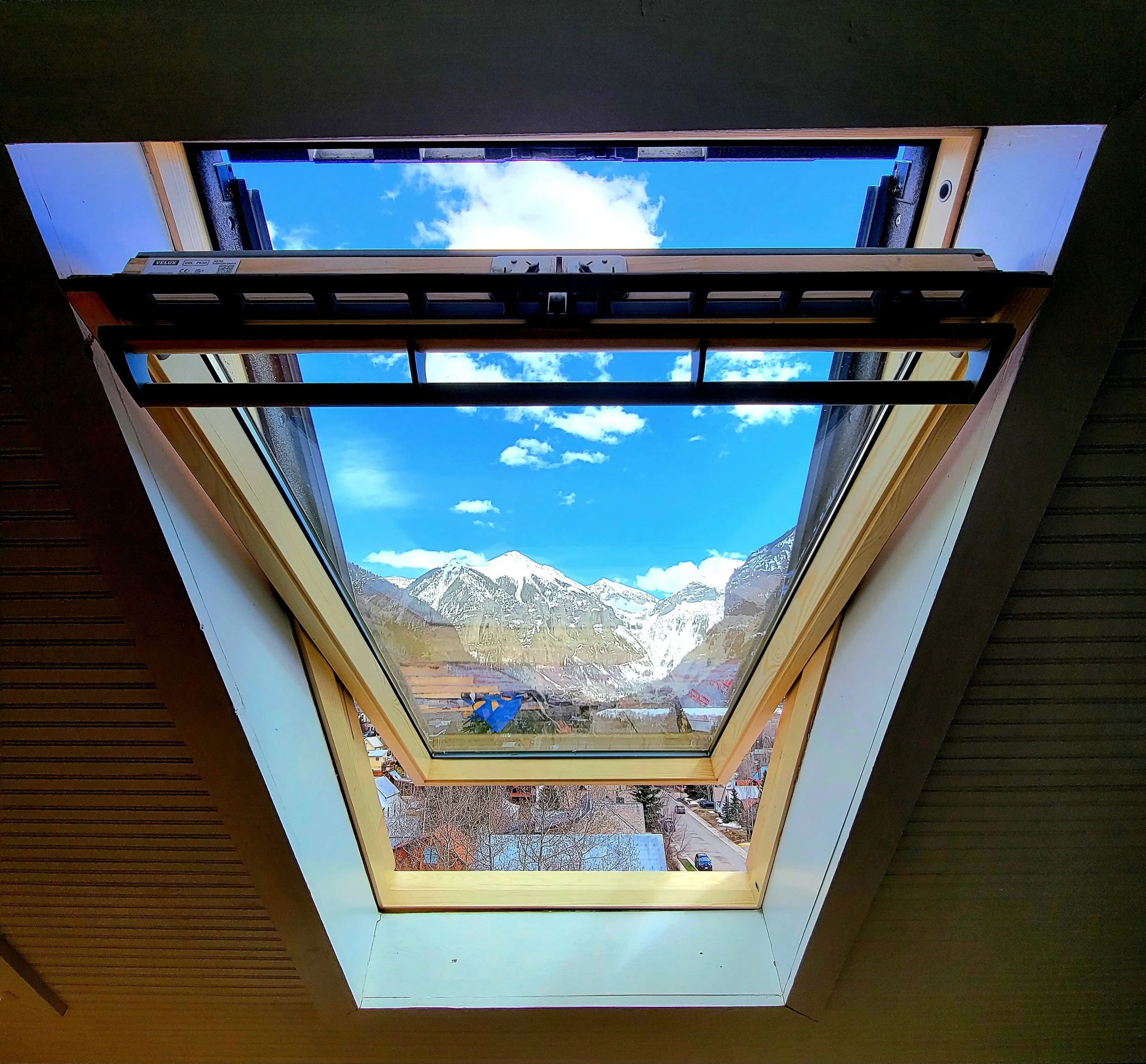 A view through an open Velux center-pivot roof window showing a clear blue sky with white clouds, the town of Telluride, and the San Juan Mountains in the background.