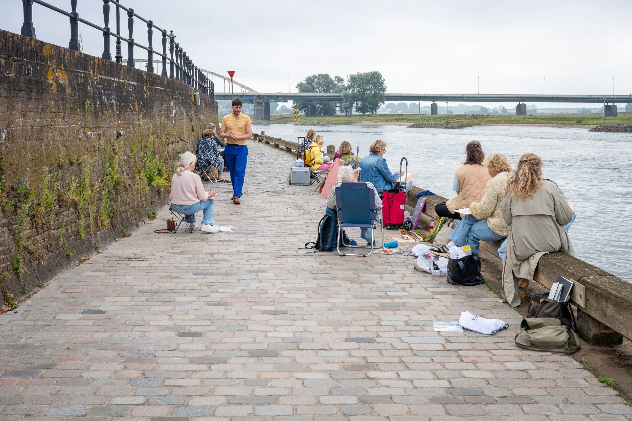 Mensen zitten langs een rivier, sommige met schepnetten voor het vissen, op een kade met een stenen muur en een houten rand, onder een bewolkte hemel.