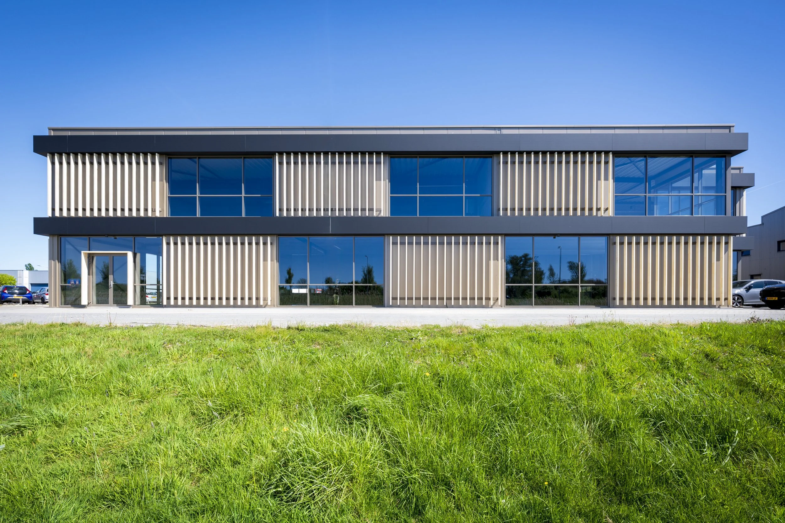 Moderne kantoorgebouw met grote glasramen en verticale houten lamellen, gelegen in een groene omgeving onder een heldere blauwe hemel.
