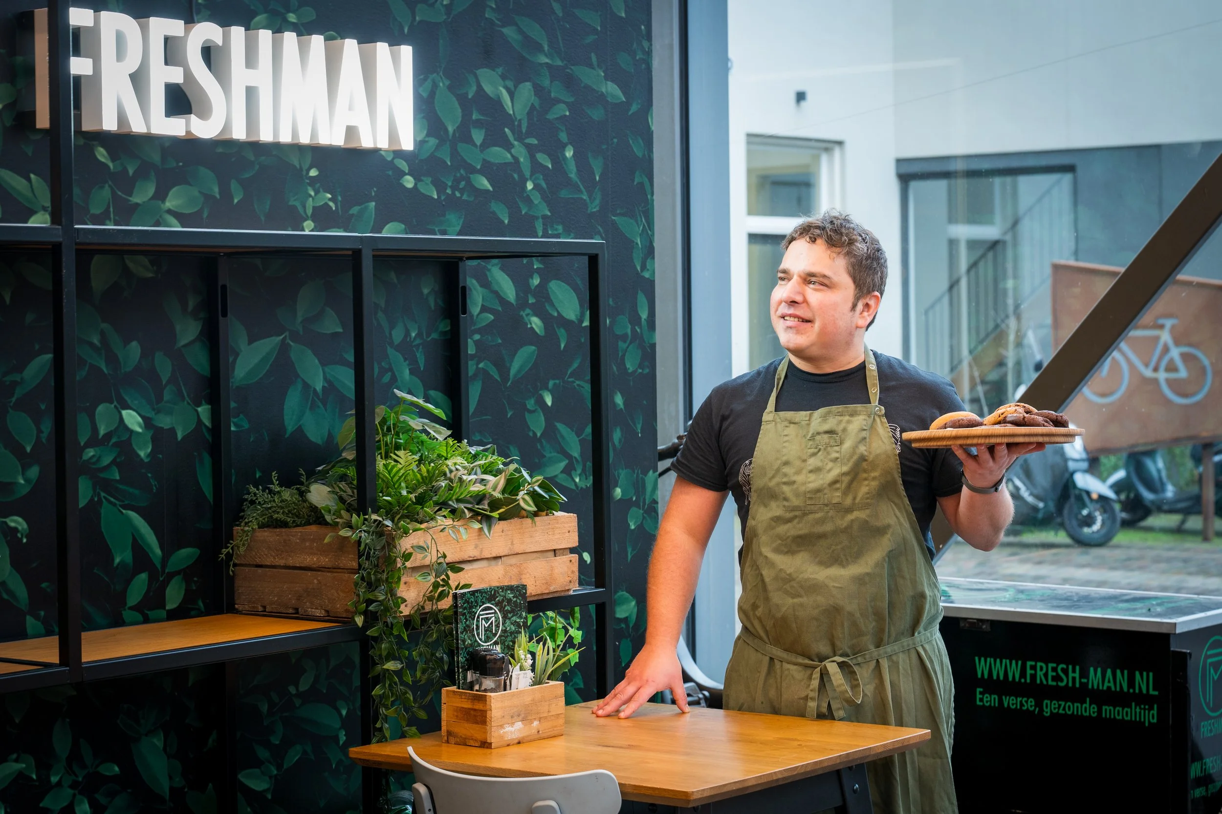 Man met een groen schort die een dienblad vol koekjes vasthoudt in een vegetarisch restaurant, met een 'FRESHMAN' teken en groene planten op de muur.
