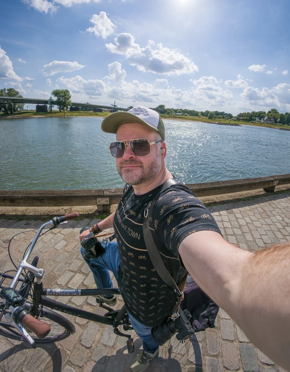 Man op de fiets neemt selfie bij water onder een heldere blauwe lucht met wolken.