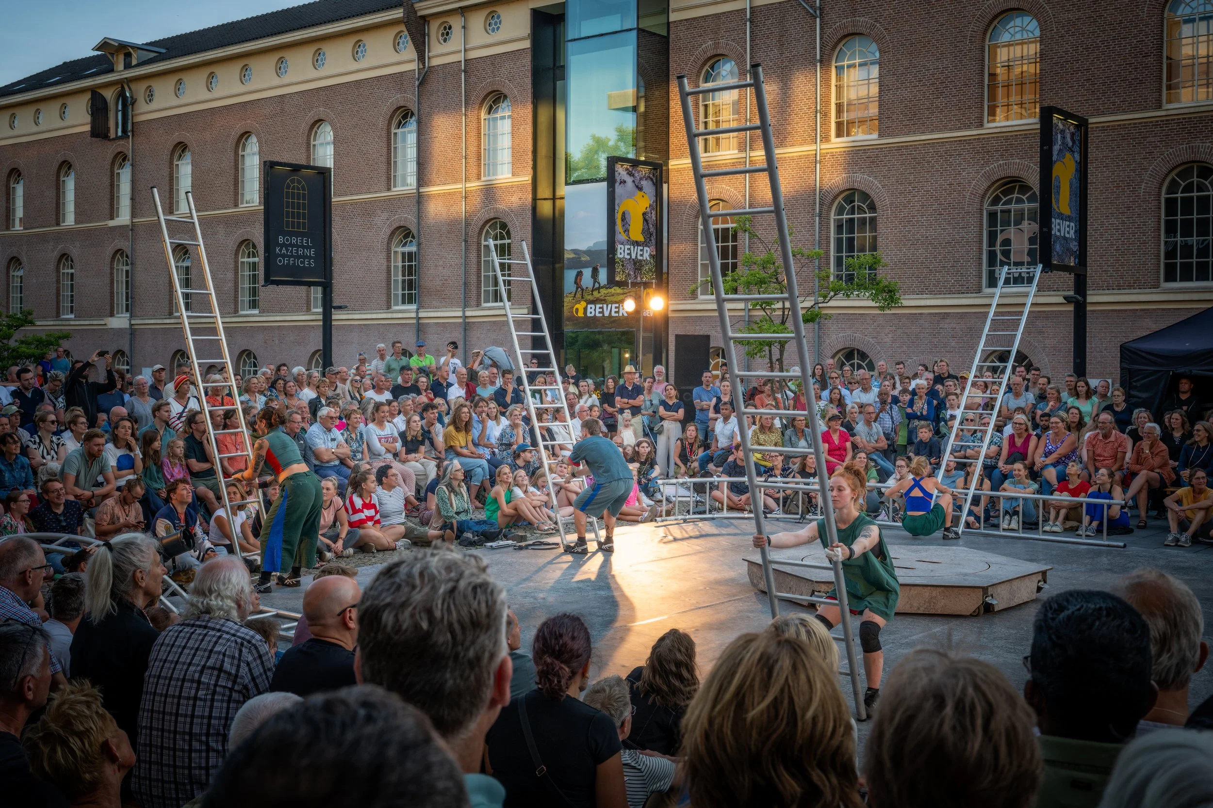 Een grote menigte mensen zit op stoelen en staat rondom een circusring, waar een artiest met groene broek en geklede haren samen met een persoon in sportkleding een act uitvoert. Op de achtergrond een gebouw met rode bakstenen en grote ramen, en enke