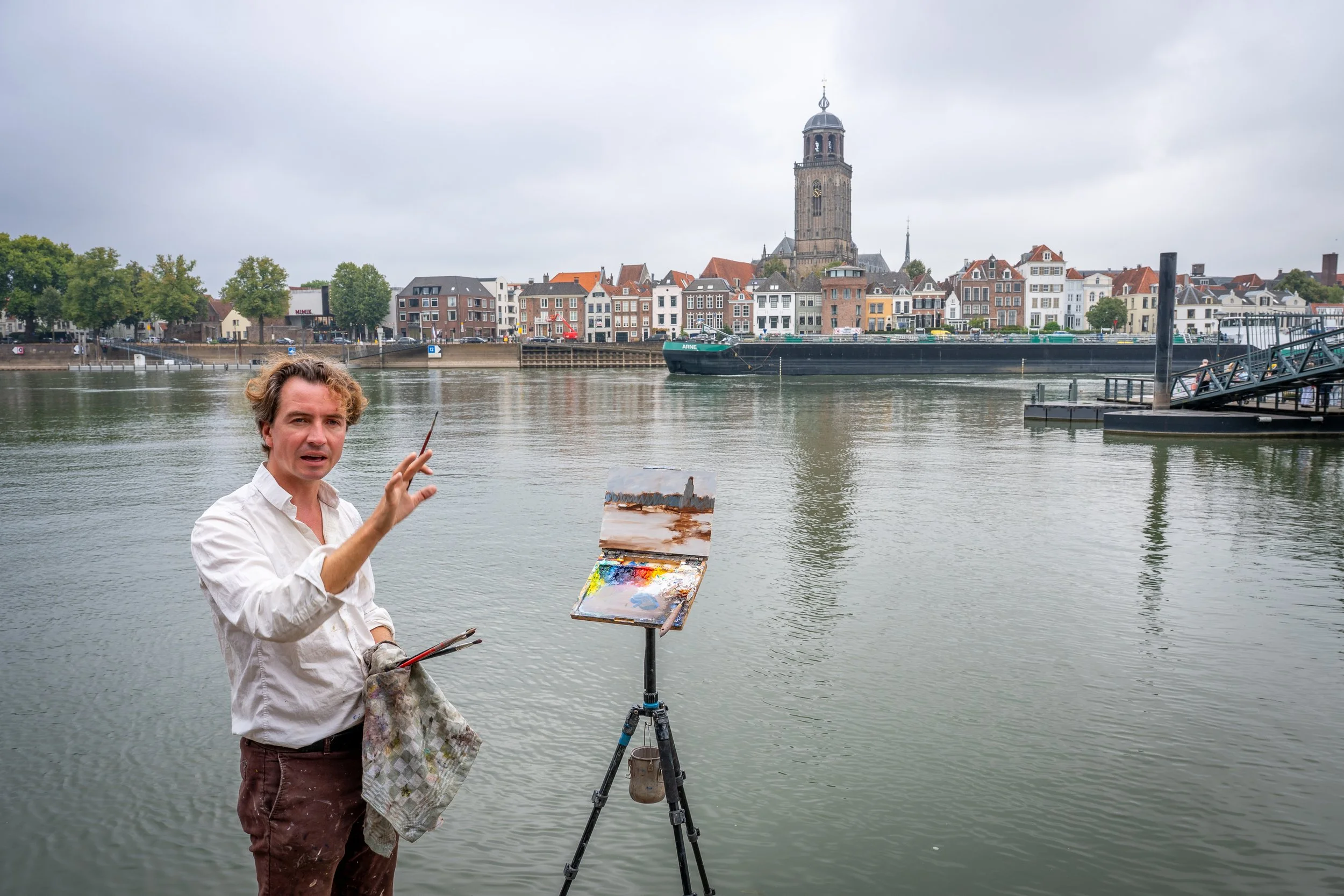 Een man met een blanke huidskleur, gekleed in een witte overhemd en bruine broek, staat bij de rivier terwijl hij schildert achter een ezel met een palet en kwasten. Op de achtergrond is een stad met kleurrijke gebouwen en een hoge kerktoren.