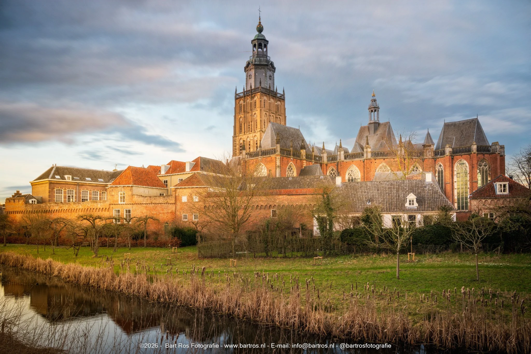 Een historische kerk met hoge toren en rode bakstenenmuren staat in een rustige omgeving, met een waterkant en grasveld voor de kerk. De lucht is deels bewolkt. Foto van Zutphen.