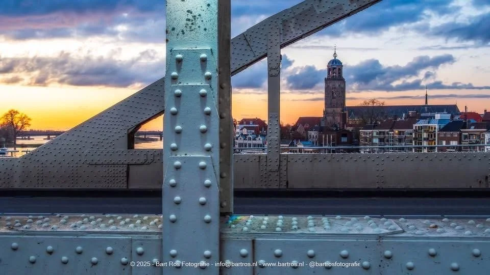 Uitzicht vanaf een brug op een stad met historische gebouwen en een kerk, tijdens zonsondergang.