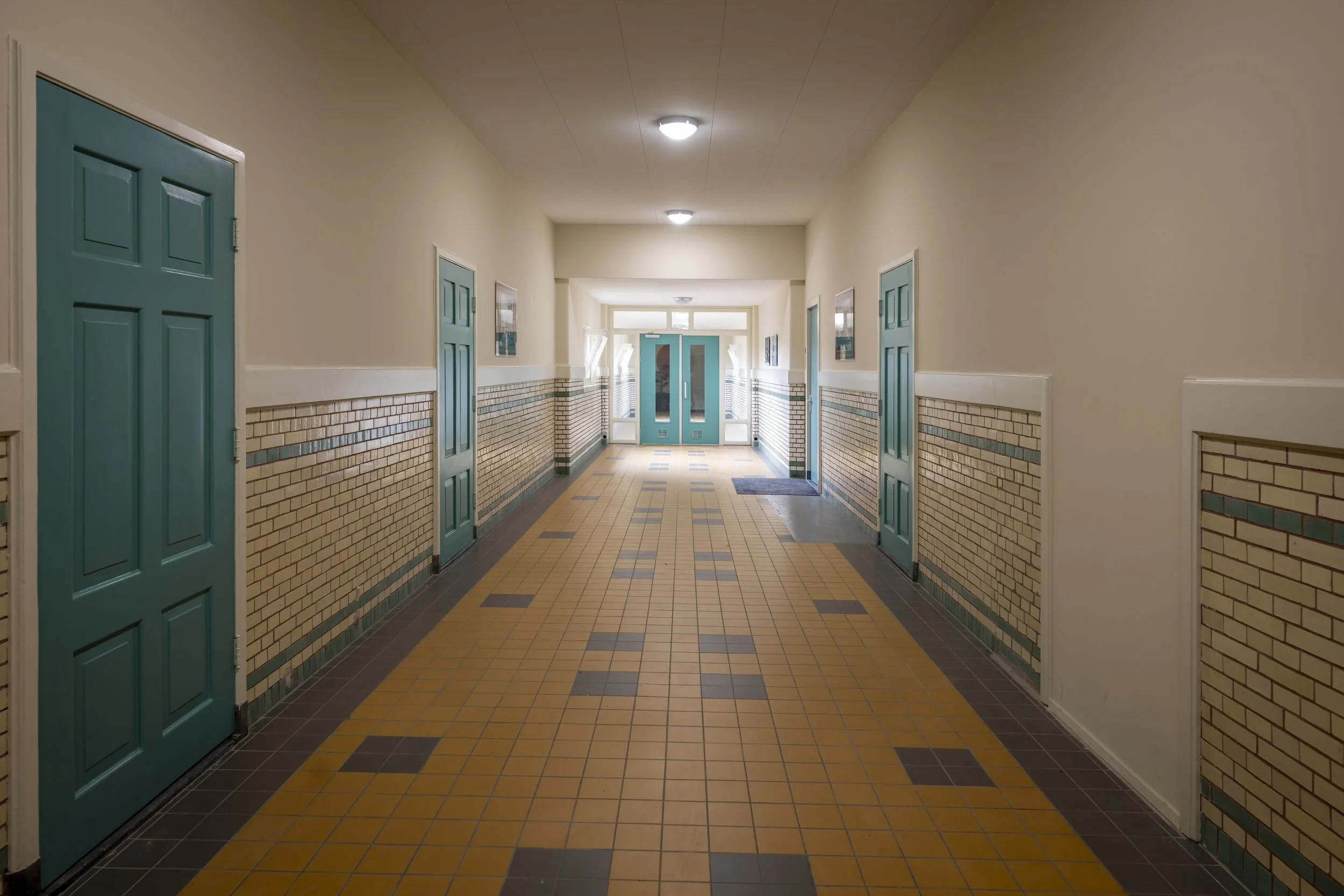Lange corridor met geel en bruin tegelvloer en blauwe deuren aan beide zijden, plafondverlichting en dubbele buitendeuren aan het einde.
