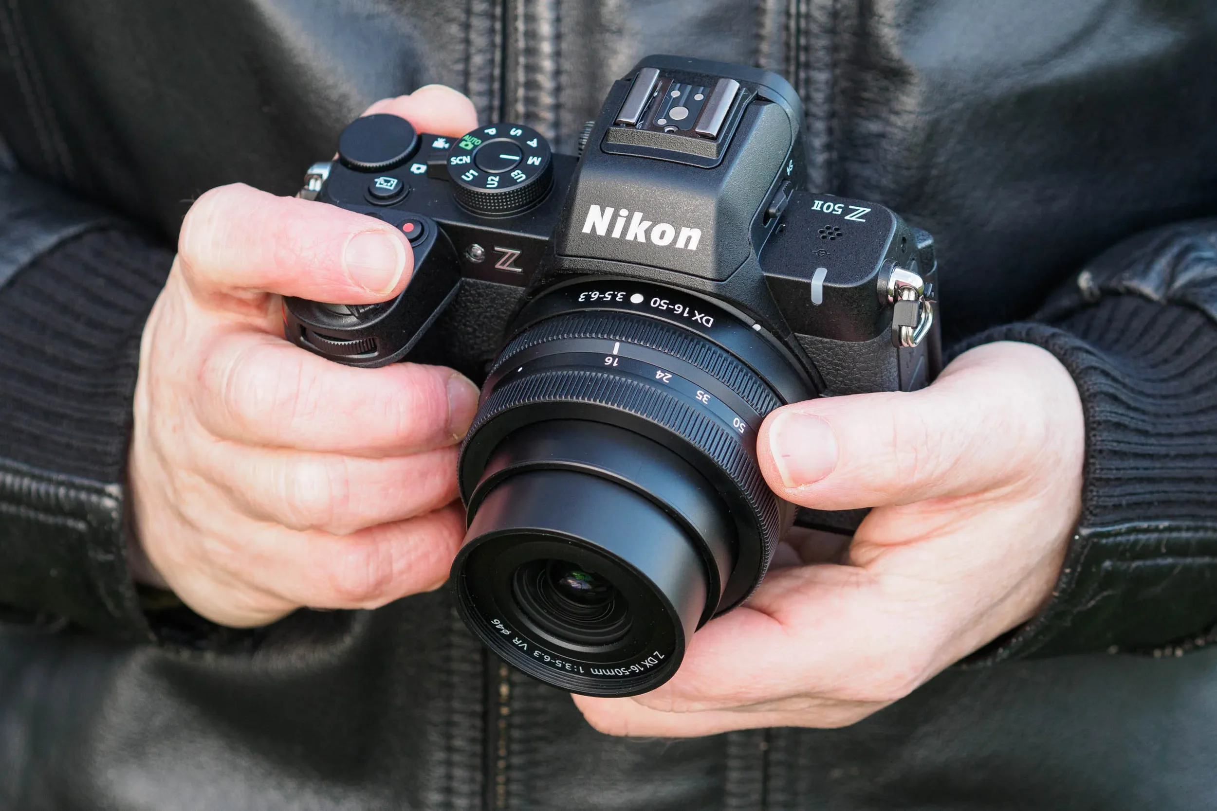 Persoon houdt een Nikon Z50ii digitale spiegelreflexcamera vast met beide handen, gekleed in een donker jasje.