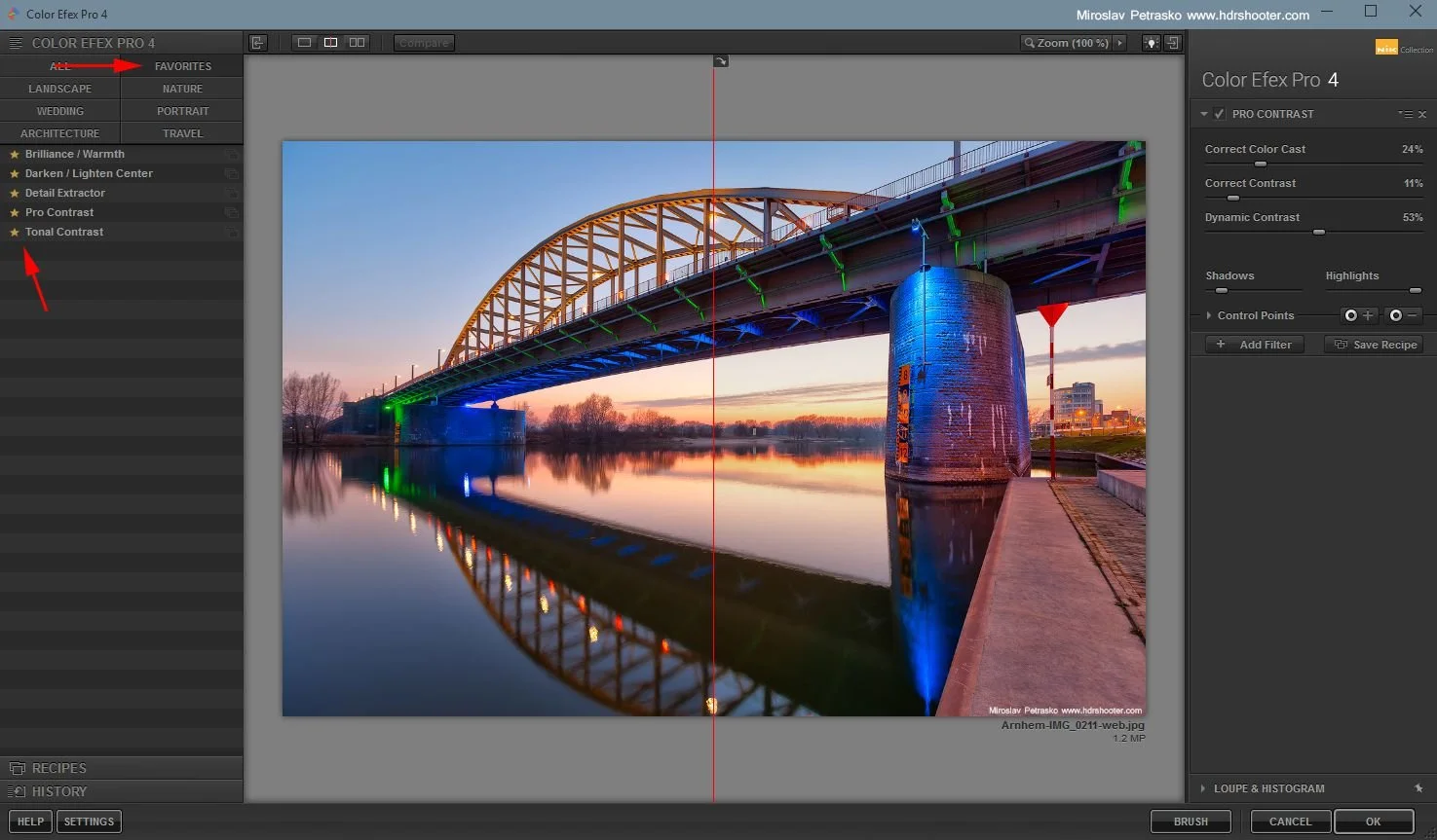 Foto van een brug over een rivier bij zonsondergang, met verlichting die het brugdek en de pylonen blauw en groen highlight, en een weerspiegeling in het water.