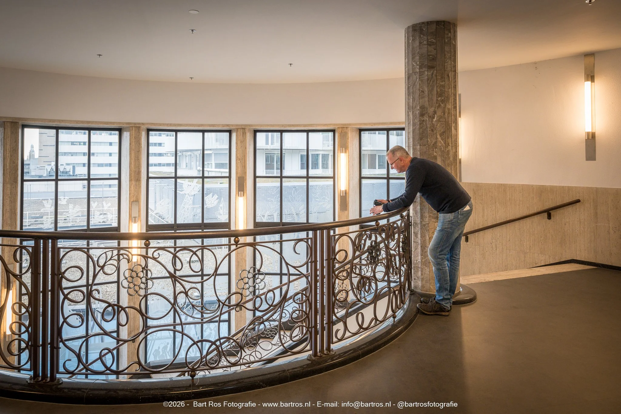 Een man in een donker shirt en spijkerbroek leunt over een decoratieve metalen balustrade in een gebouw, met grote ramen op de achtergrond die uitkijken op andere gebouwen.