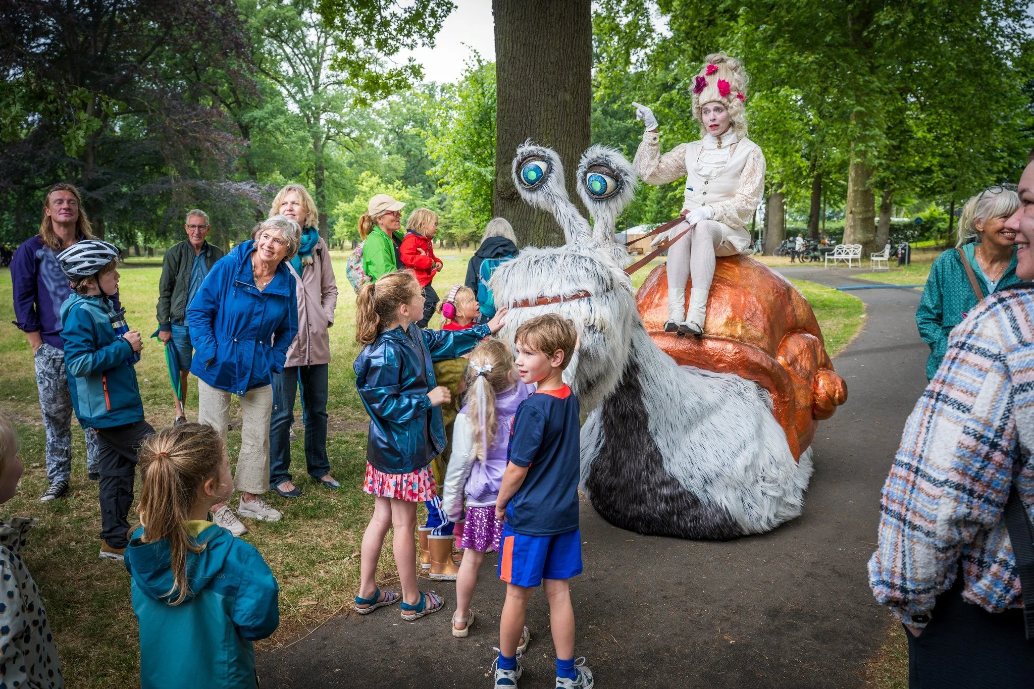 Een groep mensen, inclusief kinderen, kijkt naar een figurentheateroptreden in een park. De voorstelling wordt gedaan door een vrouw in vintage-kleding en een grote wollige figuur met grote ogen.