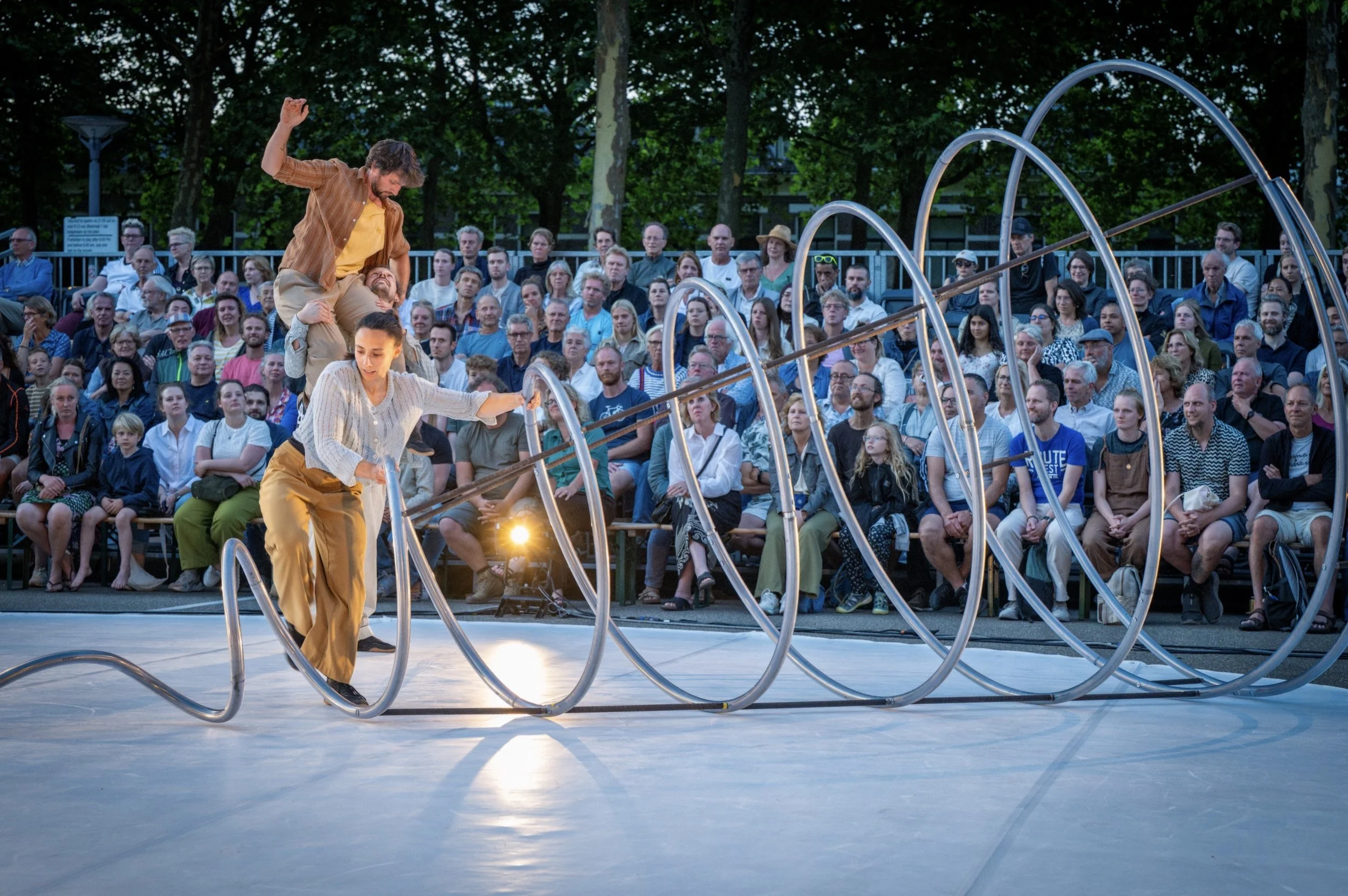 Twee artiesten uitvoeren een acrobaatkunst met metalen cirkels voor een publiek op een buitenpodium in de avond.